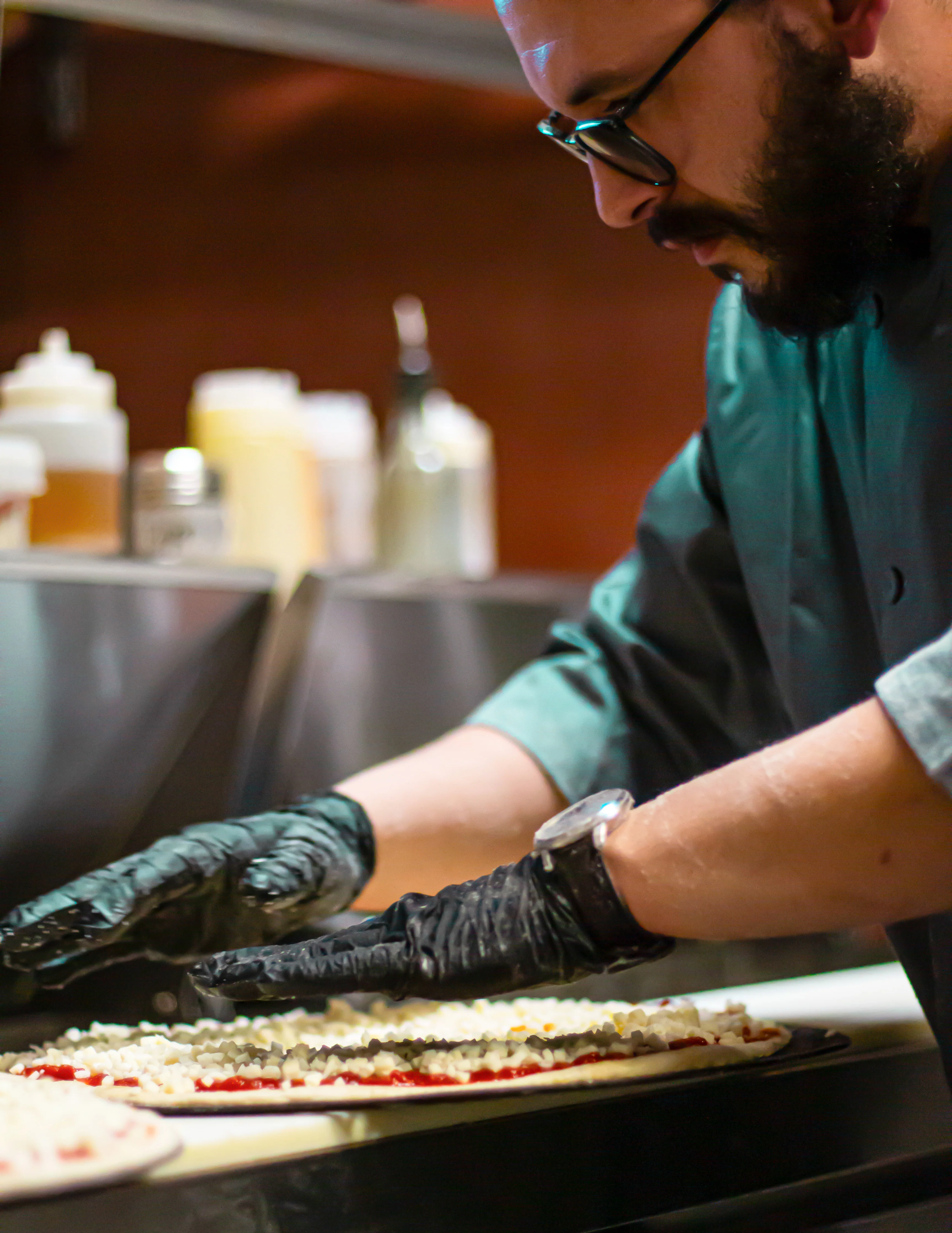A chef in a black shirt laying out toppings on a pizza base in a busy kitchen.