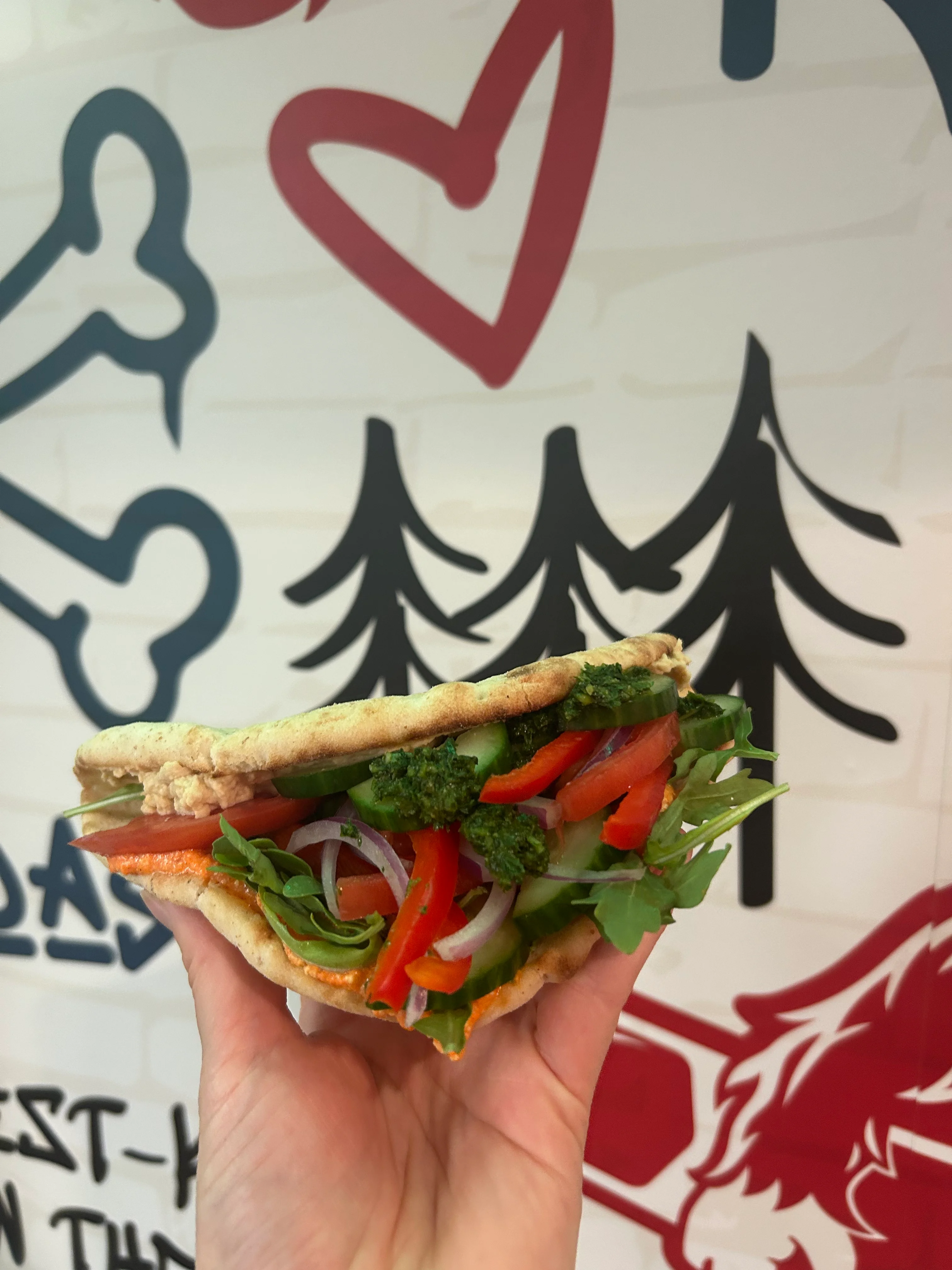 A hand presenting a stuffed flatbread filled with colorful vegetables, including bell peppers and greens, displayed against a creative wall design.