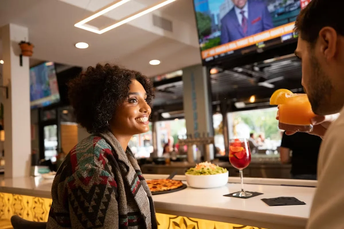 A woman smiling at a bar with drinks and food in a lively restaurant setting.