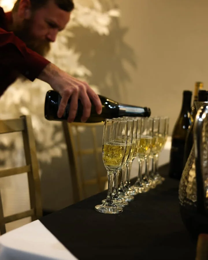A man with a beard and maroon shirt carefully pours sparkling wine into a row of champagne flutes set on a black tablecloth. The golden chairs and soft lighting in the background create an elegant ambiance, suggesting a celebratory or upscale event.