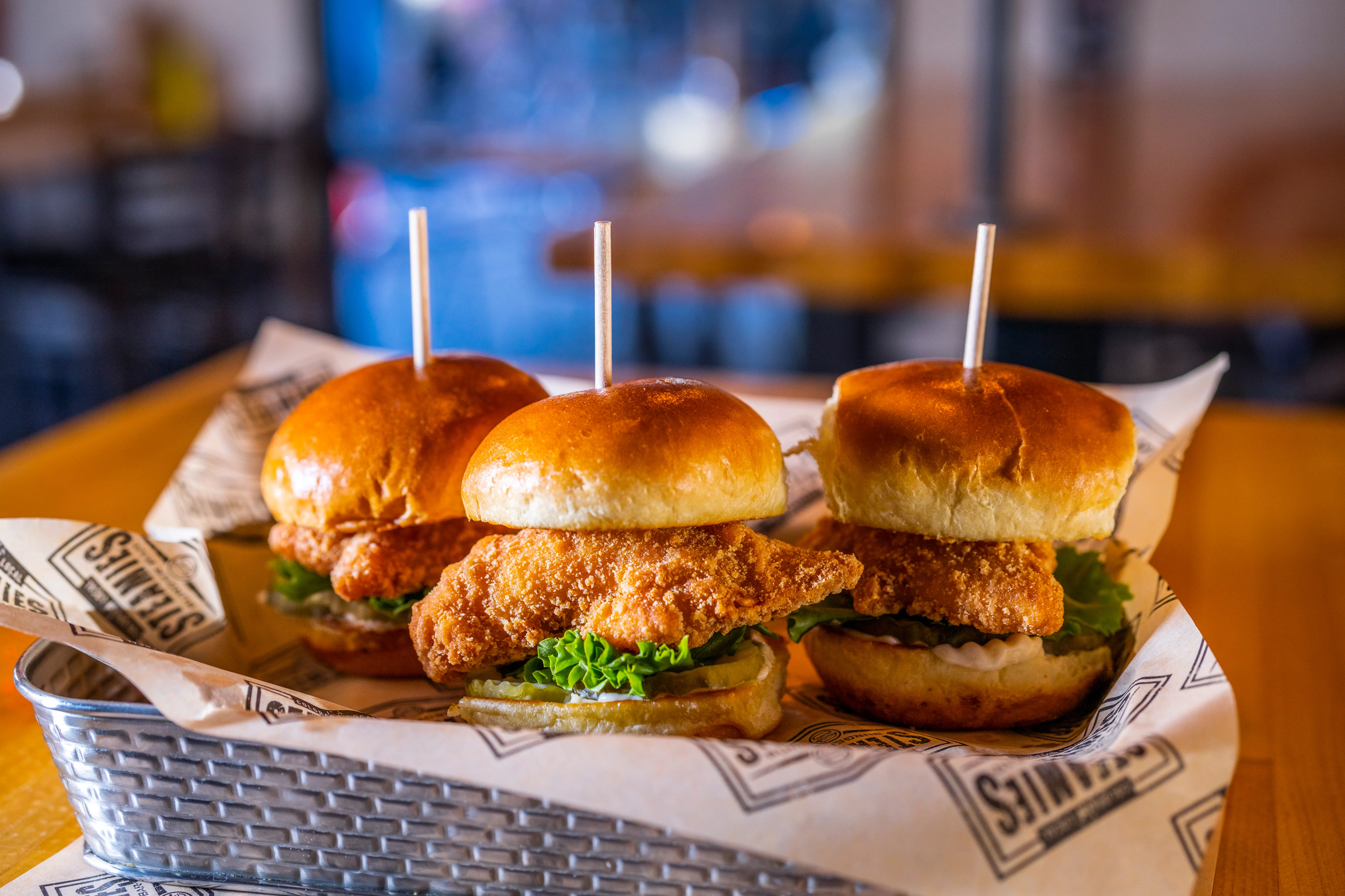 A basket of three mini chicken burgers, each topped with lettuce and mayonnaise. They are served on a branded paper liner.