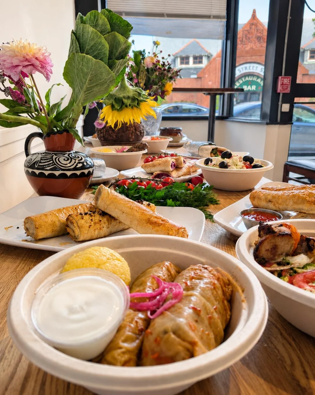 An array of vibrant dishes on a table, featuring rolled items, salads, and garnishes, accompanied by flowers and a view through the window.