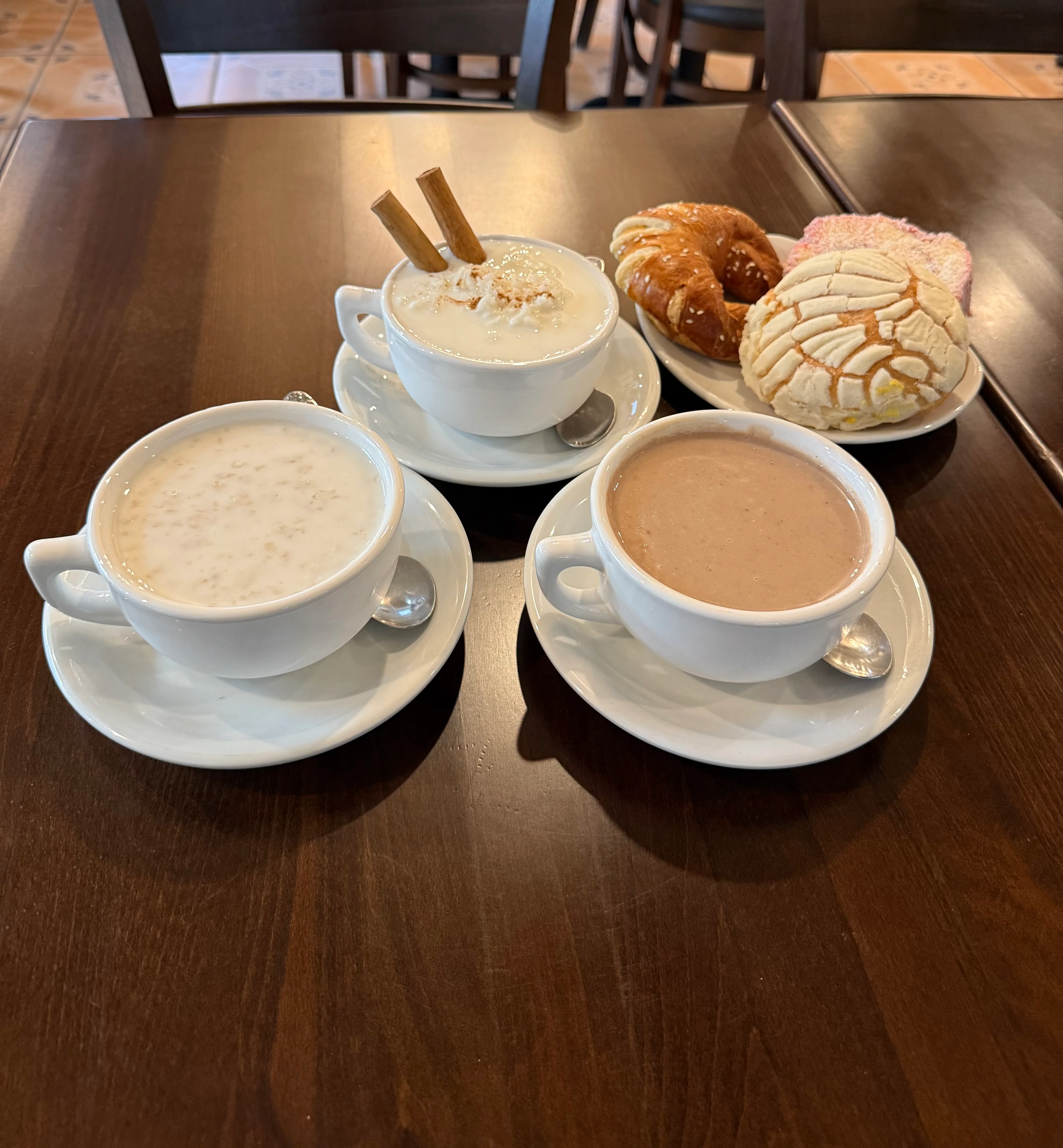 Three cups of beverages served with a selection of pastries, including a croissant and cookies, on a wooden table.