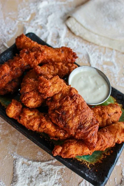 A serving of crispy fried chicken strips accompanied by a side of white dipping sauce on a dark plate.