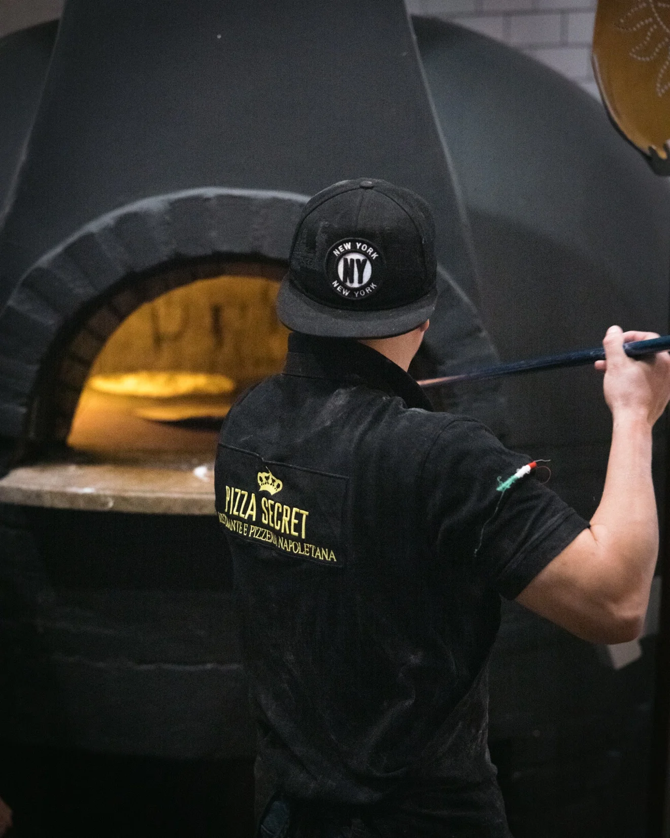 Chef at the pizza oven, back view showing Pizza Secret shirt