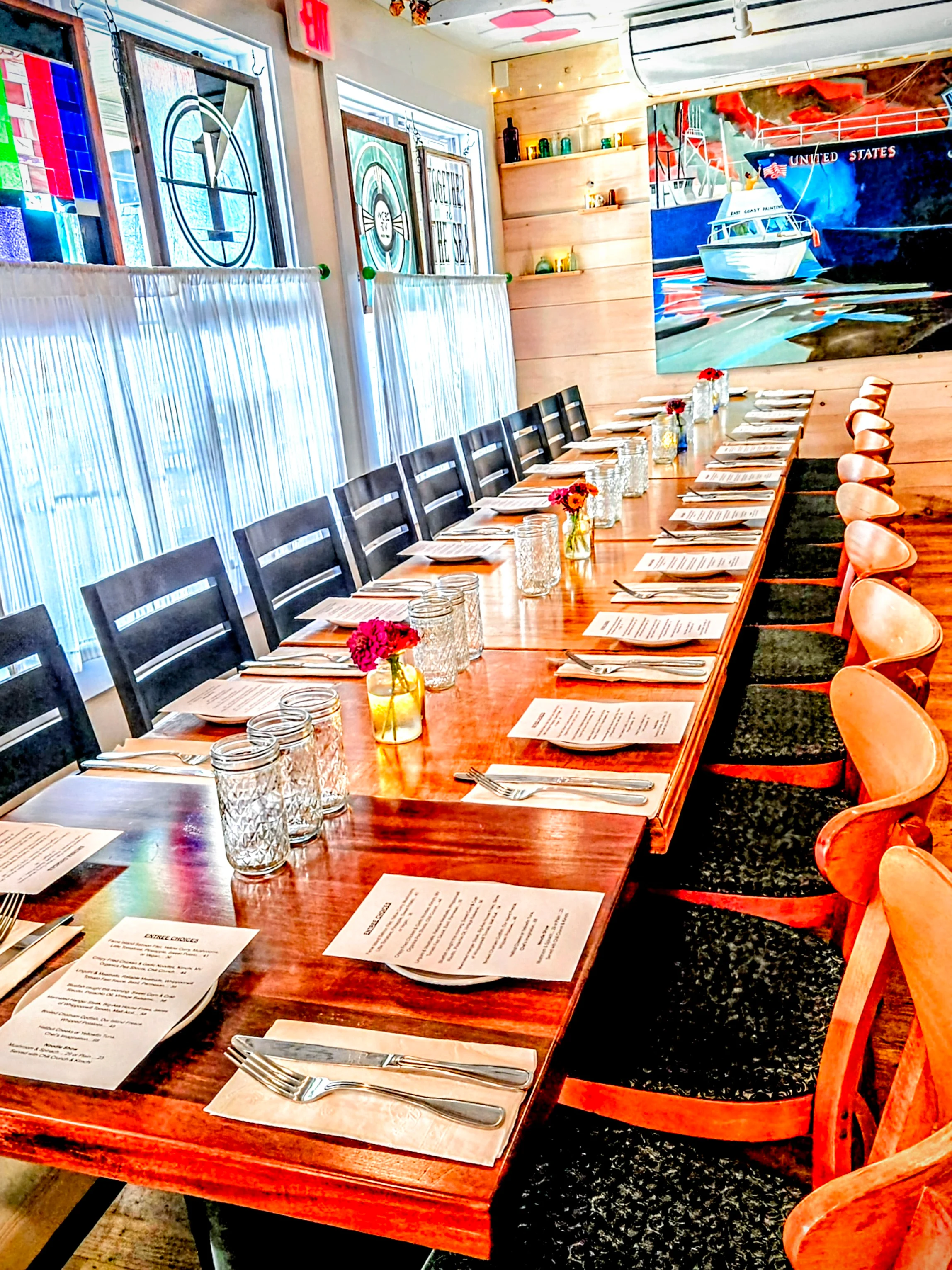 A long dining table set with menus, silverware, and flowers, illuminated by natural light from large windows.