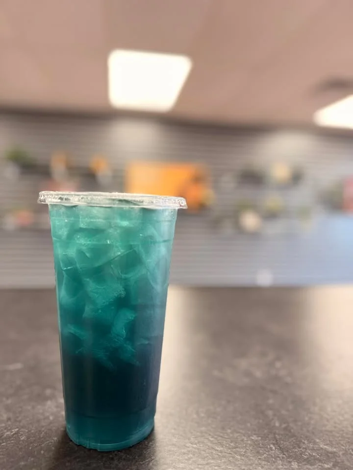 A layered teal drink with ice, placed on a dark surface in a well-lit interior.