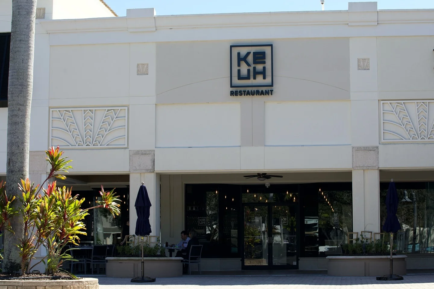 The bright exterior of Ke-uH Boca Raton during the day, highlighting the main square logo on a white building façade above a covered patio area with glass doors and closed blue umbrellas.