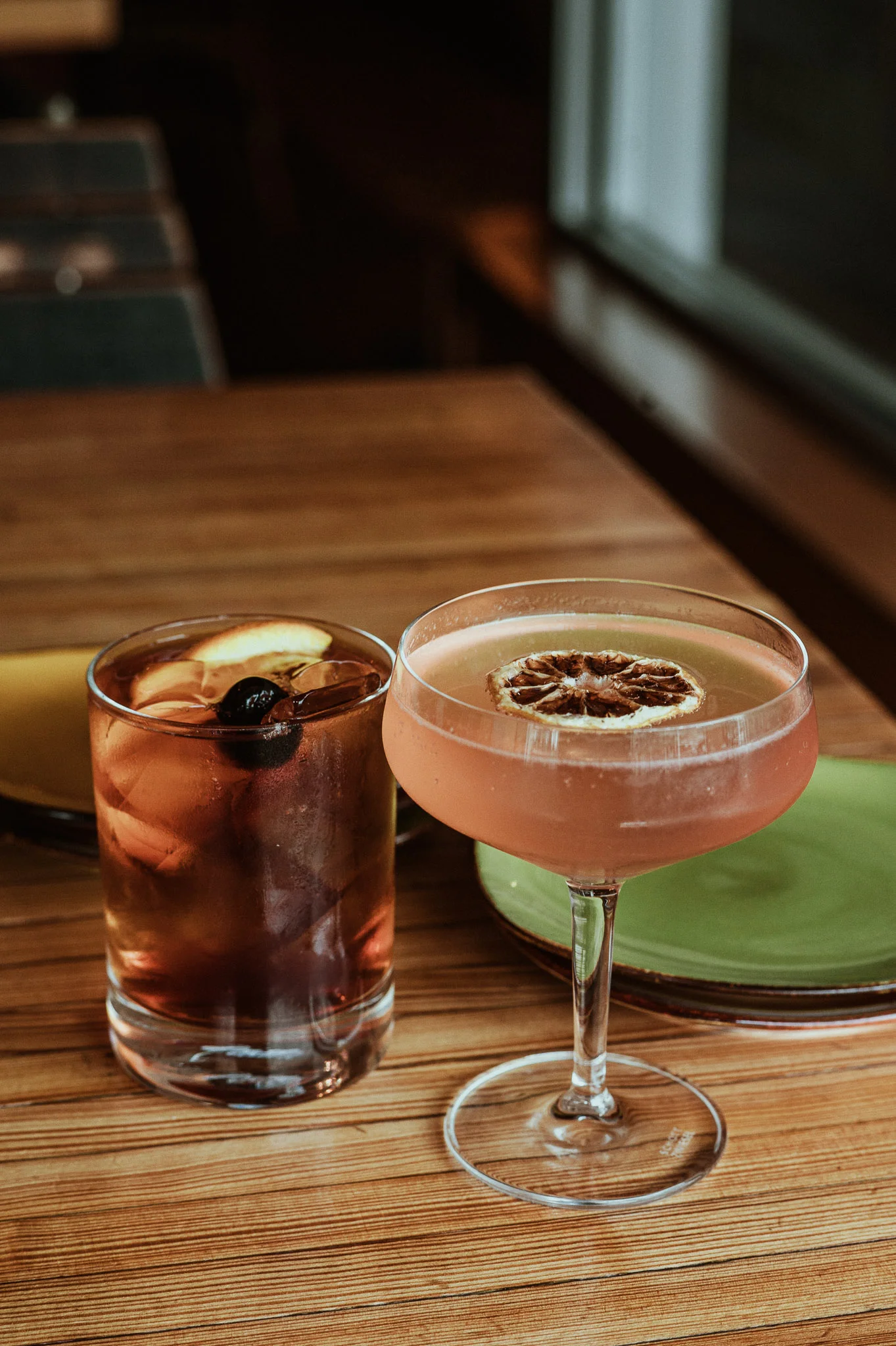 Two cocktails on a wooden table: one in a rocks glass, one in a coupe glass.