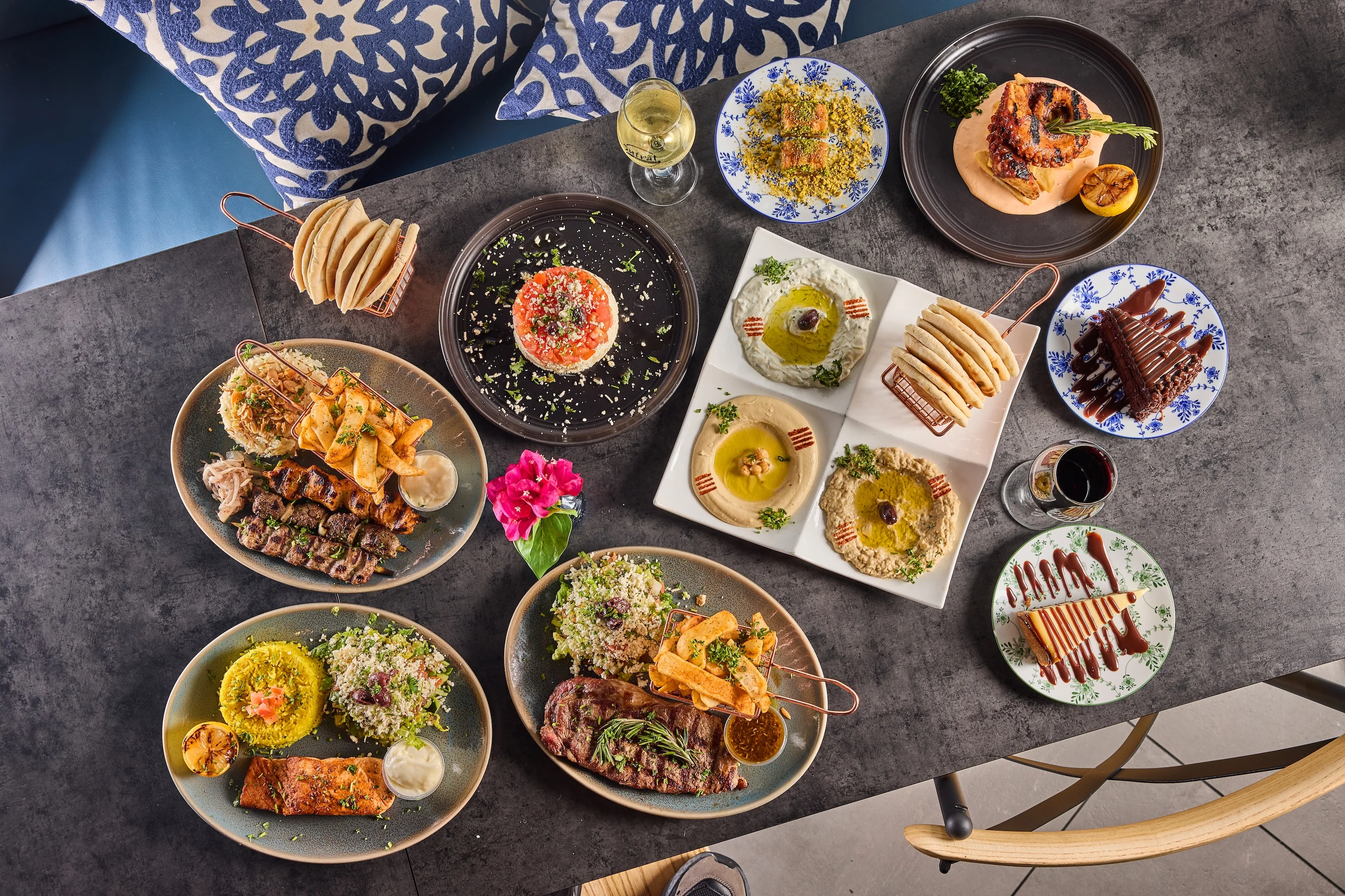 An overhead view of a table filled with colorful Middle Eastern dishes, including hummus, grilled meats, and desserts, accompanied by drinks. The scene is vibrant and inviting, set against a dark table with blue-patterned cushions.