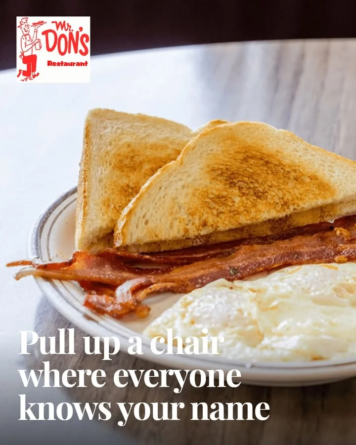 A plate with crispy bacon, toast, and eggs served on a white dish. The setting suggests a cozy dining atmosphere.