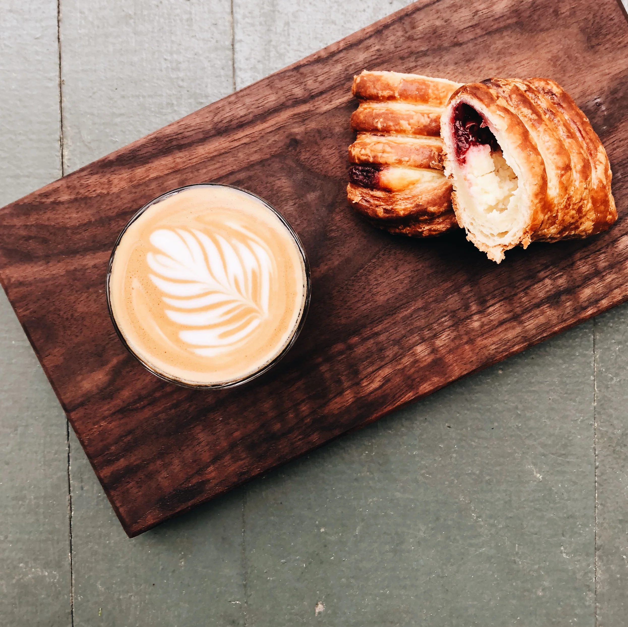 A cup of latte with intricate foam art sits next to a flaky pastry filled with fruit and cream on a wooden board.