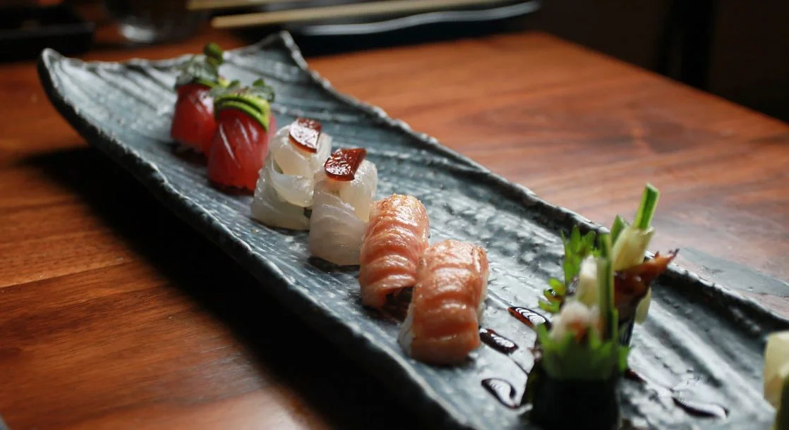 An assortment of premium nigiri and gunkan sushi arranged in pairs along a long, textured dark grey ceramic plate, featuring tuna with avocado, white fish with a dark glaze, seared salmon, and eel rolls garnished with green scallions on a wooden table.