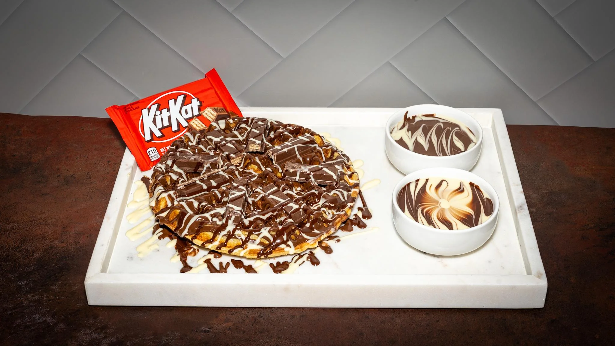 A dessert pizza topped with pieces of Kit Kat bars, drizzled with chocolate, and accompanied by two bowls of dipping sauce.
