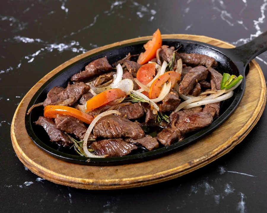 Sizzling beef served on a cast iron plate with onions, peppers, and garnishes.