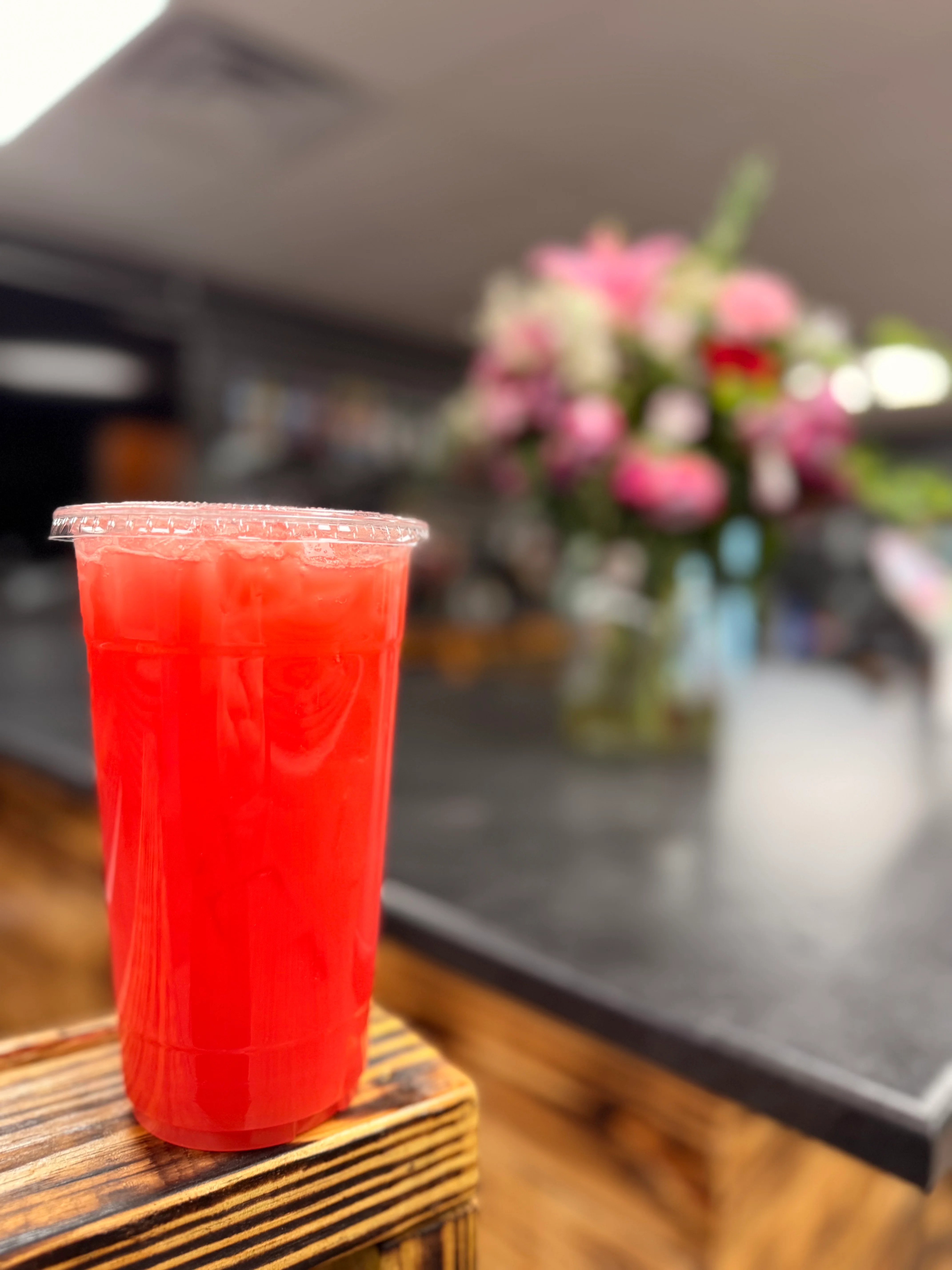 A vibrant red beverage served in a clear cup filled with ice, placed on a wooden surface, with blurred floral arrangements in the background.
