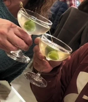 A close-up of a hand pouring a drink from a tap into a glass at a bar, with another hand visible in the background.