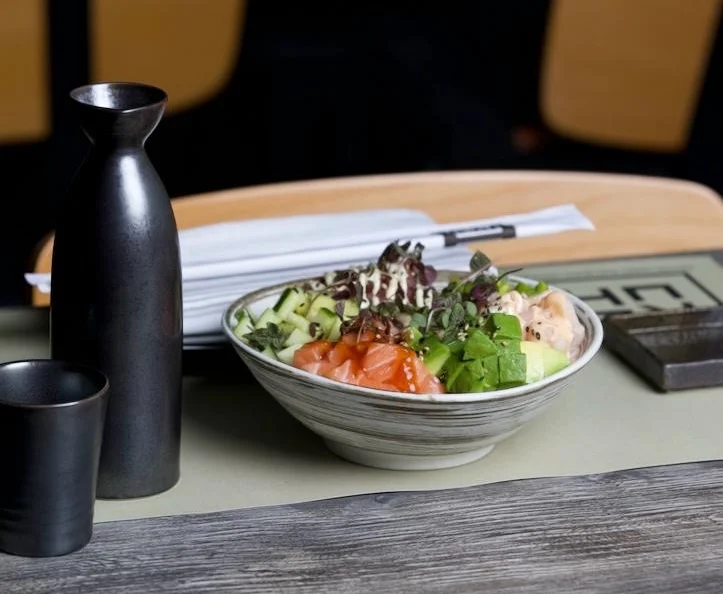 A fresh poké bowl filled with sections of diced salmon, tuna, cubed avocado, and chopped cucumber, topped with microgreens and creamy sauce, served on a restaurant table next to a dark ceramic sake carafe and matching cup.