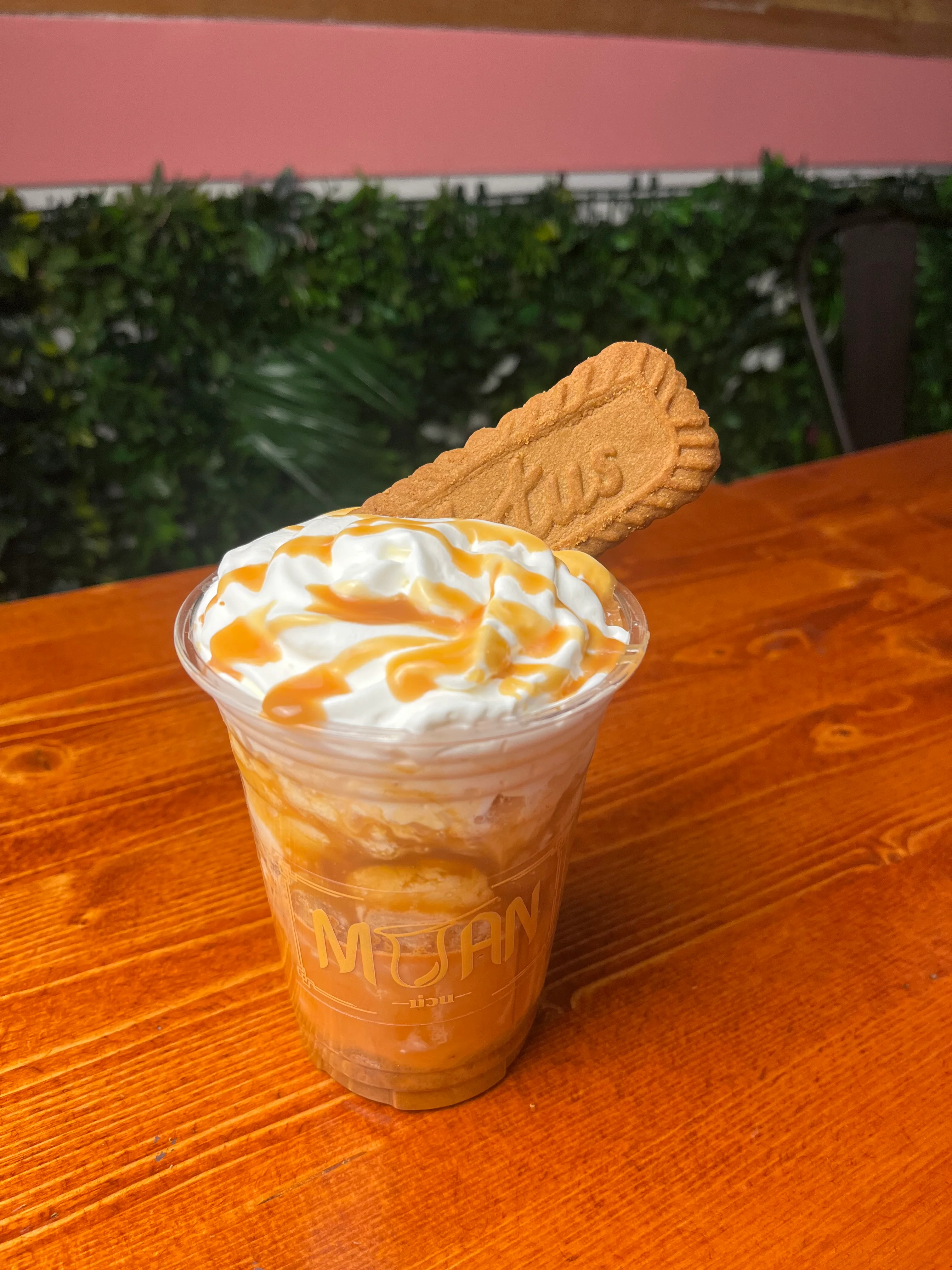 A close-up of a dessert drink topped with whipped cream, caramel drizzle, and a biscuit on a wooden table.