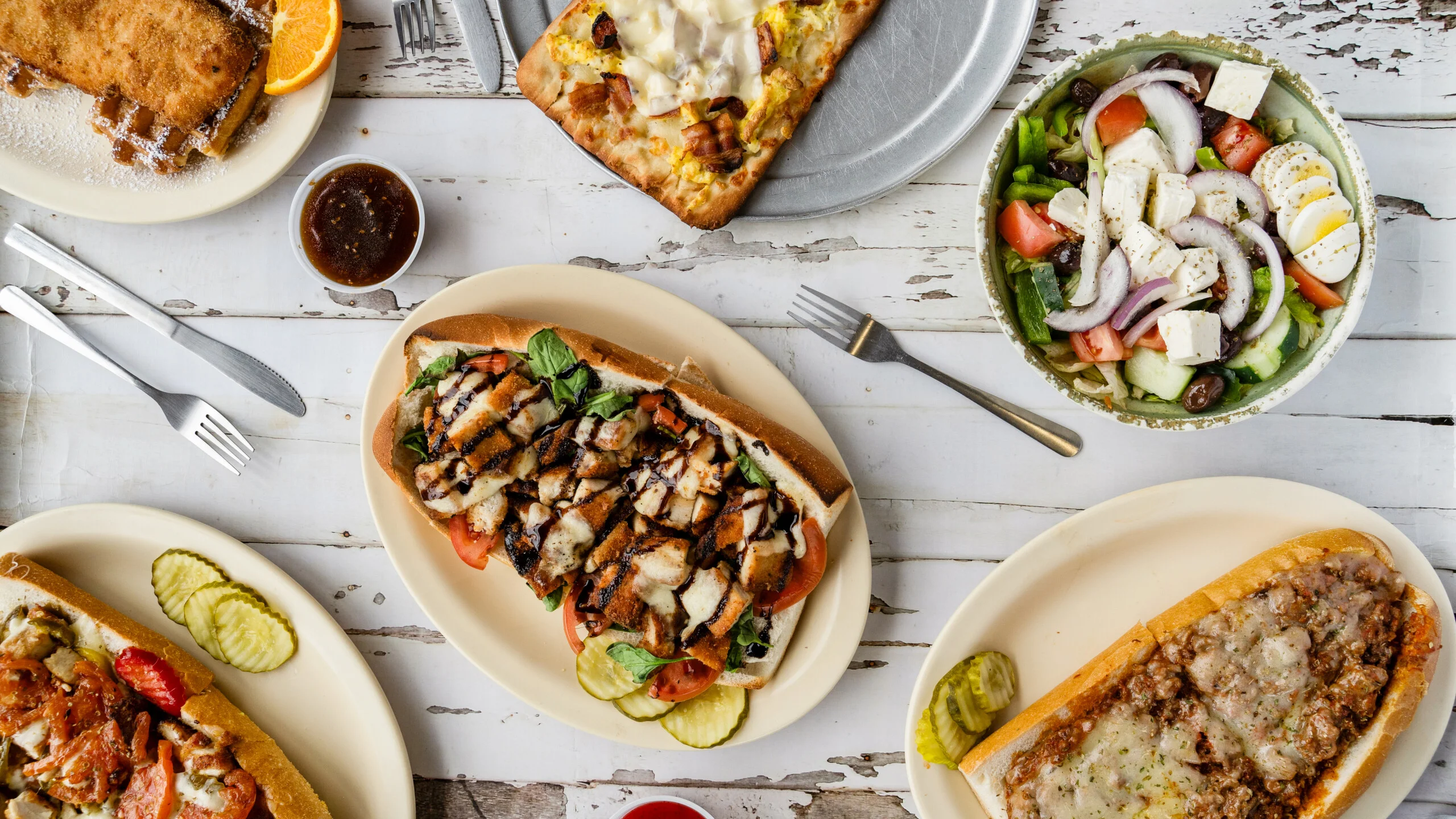 Overhead view of Steak Out cheesesteaks, hoagies, and fresh salads on white wooden table with pickles and dipping sauce.