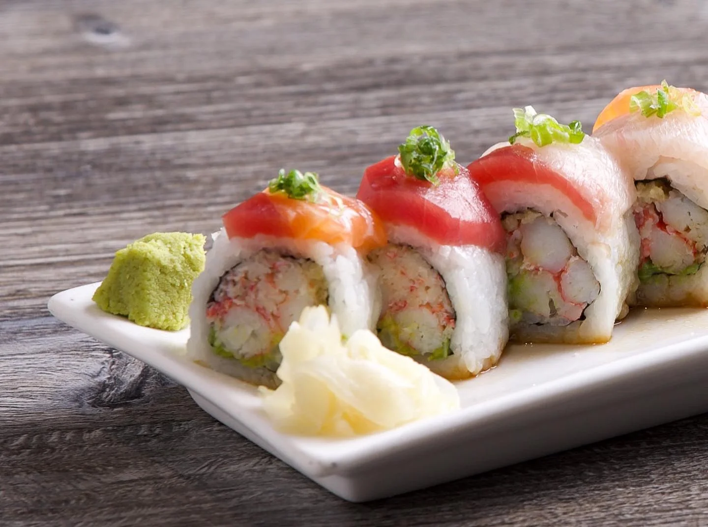A row of sliced rainbow sushi rolls filled with crab salad and avocado, topped with assorted fresh fish including salmon and tuna, and garnished with chopped green onions, served on a white square plate with a dollop of wasabi and pickled ginger on a rustic wooden table.