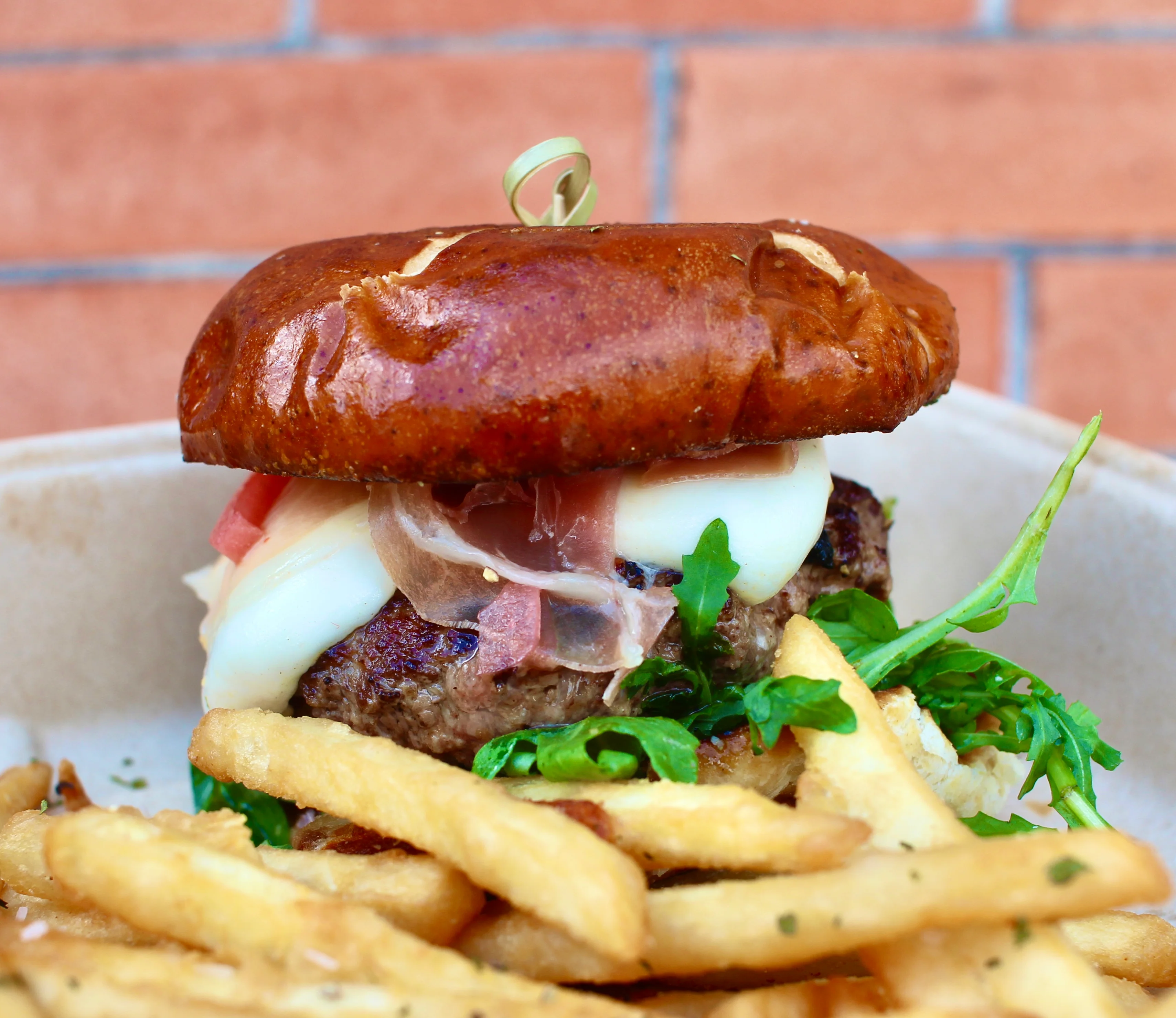A gourmet burger served on a pretzel bun, garnished with cheese, prosciutto, and greens, accompanied by crispy fries.