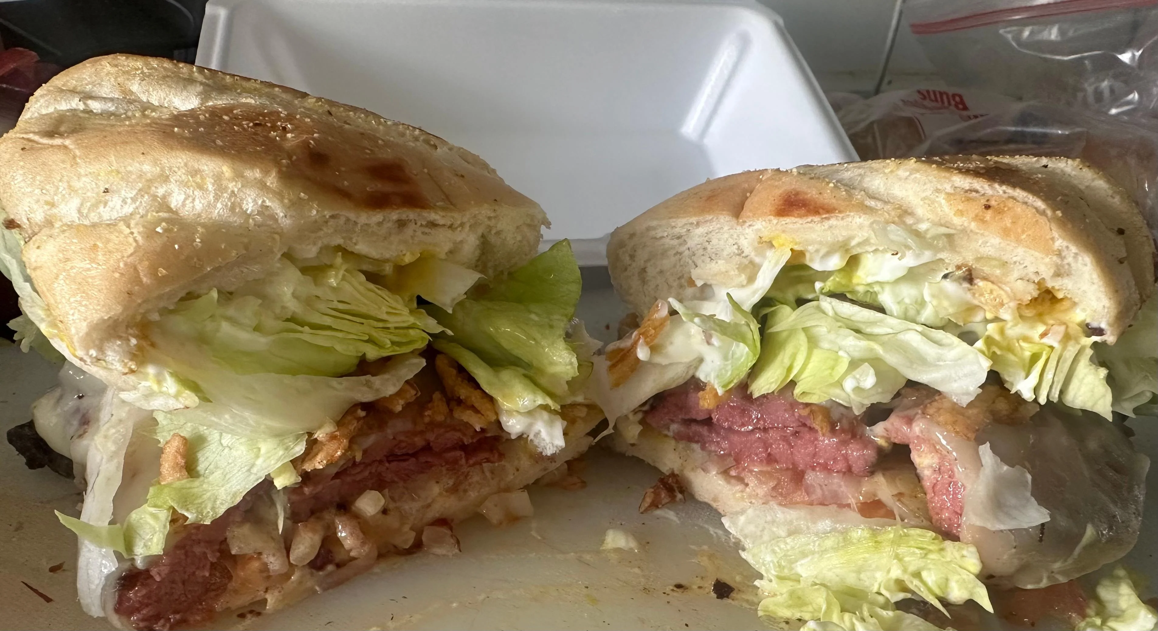 A large sandwich cut in half, filled with leafy greens, meats, and toppings. The sandwich is placed on a white takeout container.