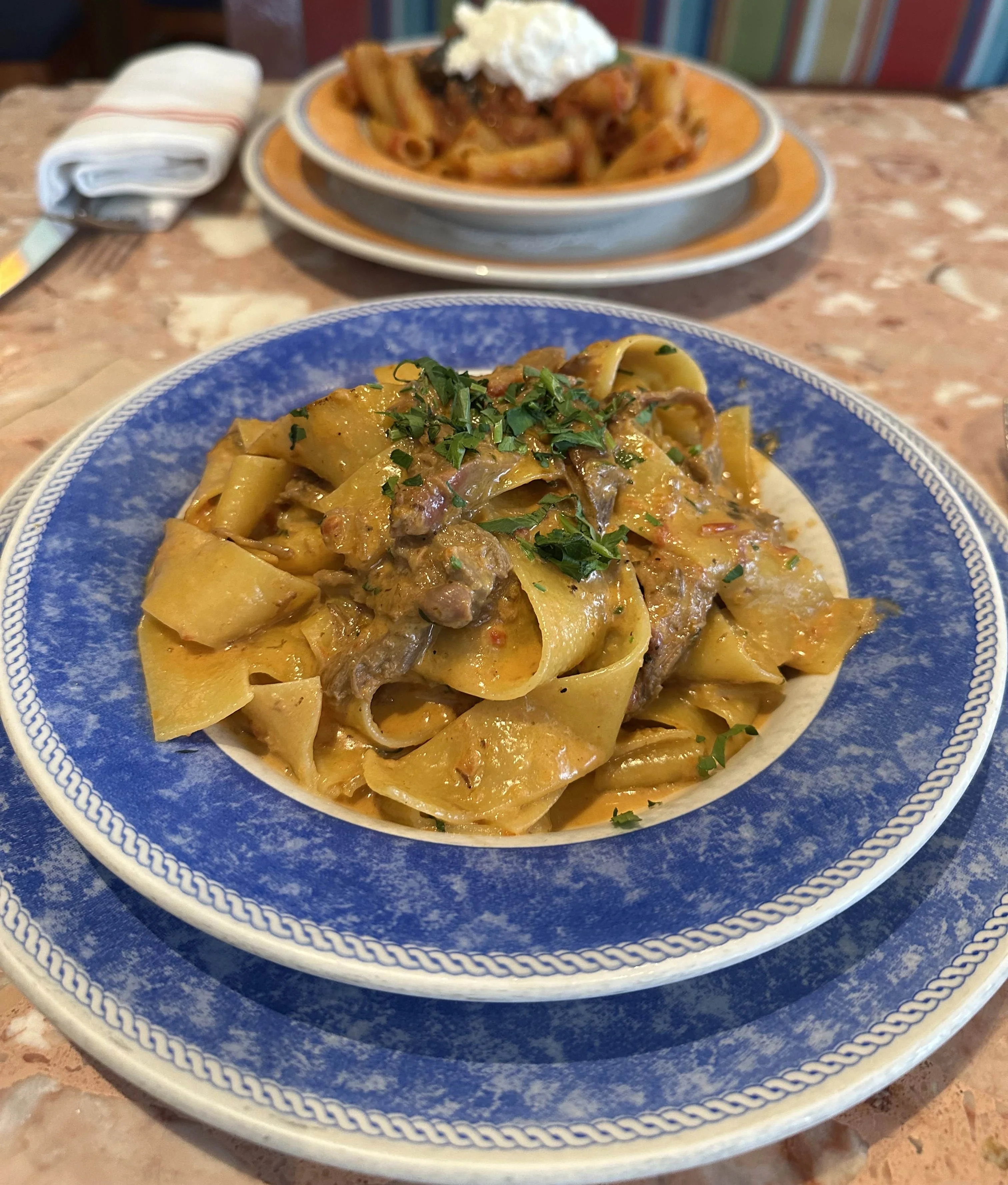 A plate of pasta served with a rich sauce and garnished with herbs. In the background, another pasta dish is topped with a creamy garnish.