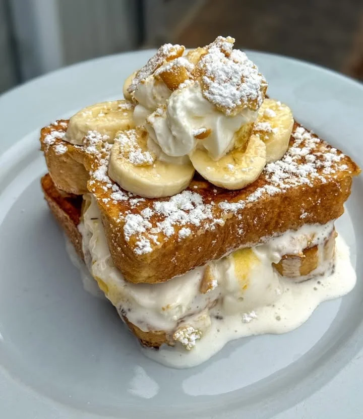 A decadent dish featuring layers of French toast, bananas, whipped cream, and powdered sugar.