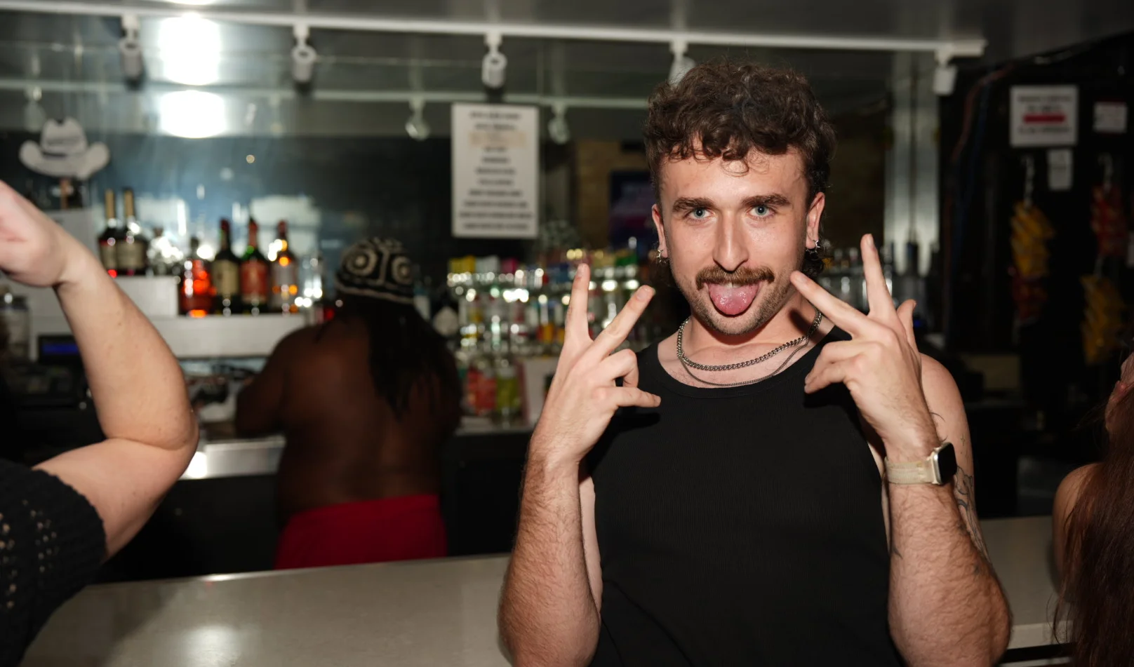 A person making a playful gesture at a bar with drinks in the background.