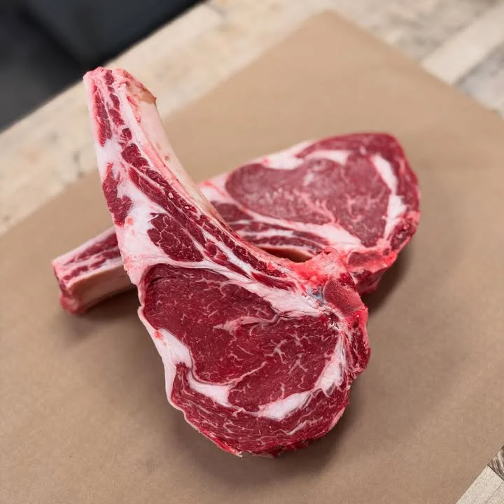 Two large cuts of ribeye steak displayed on brown paper, highlighting their marbling and quality.