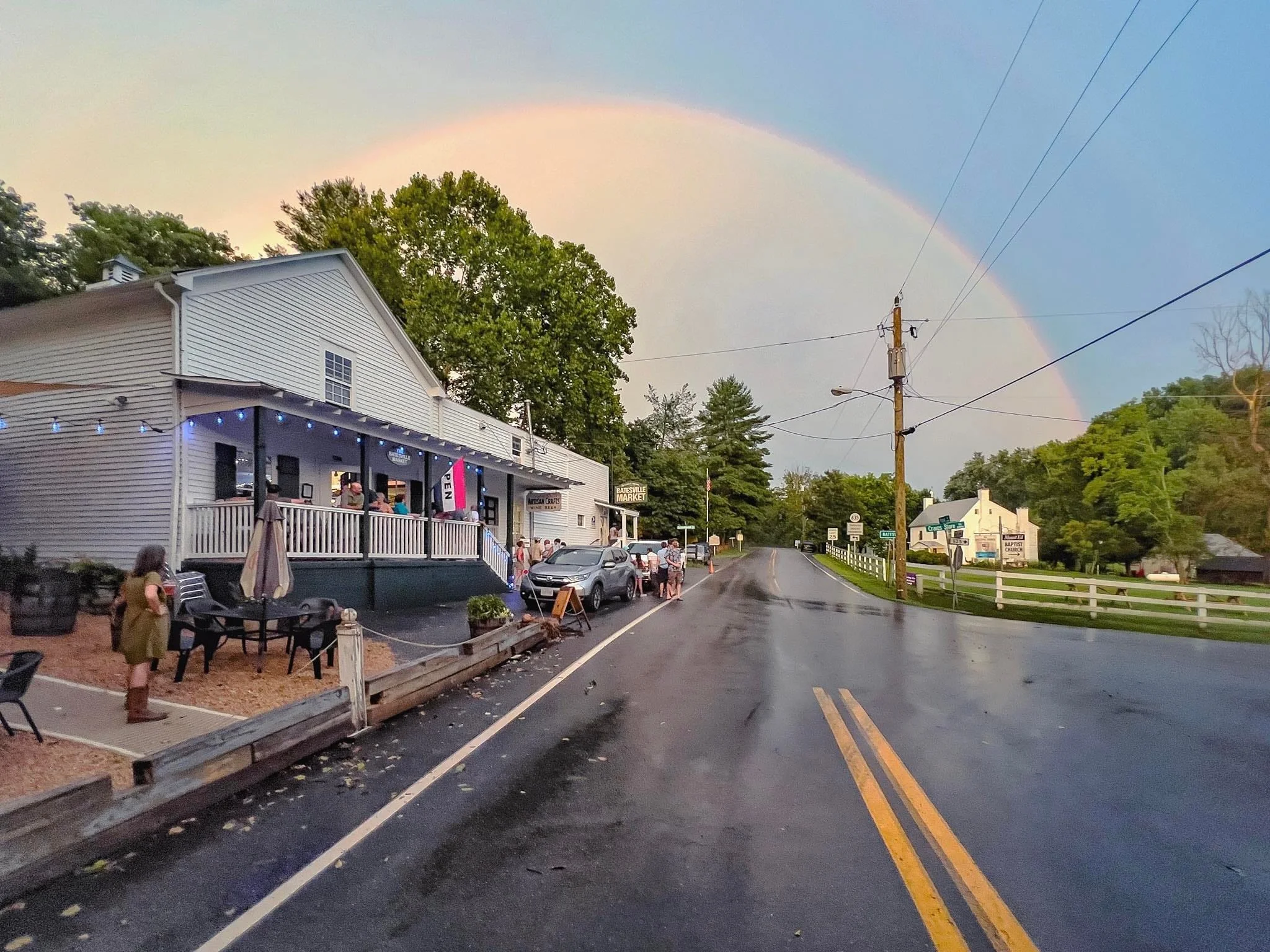 A picturesque street scene featuring a charming building under a rainbow, with people socializing outside on a peaceful rainy day.