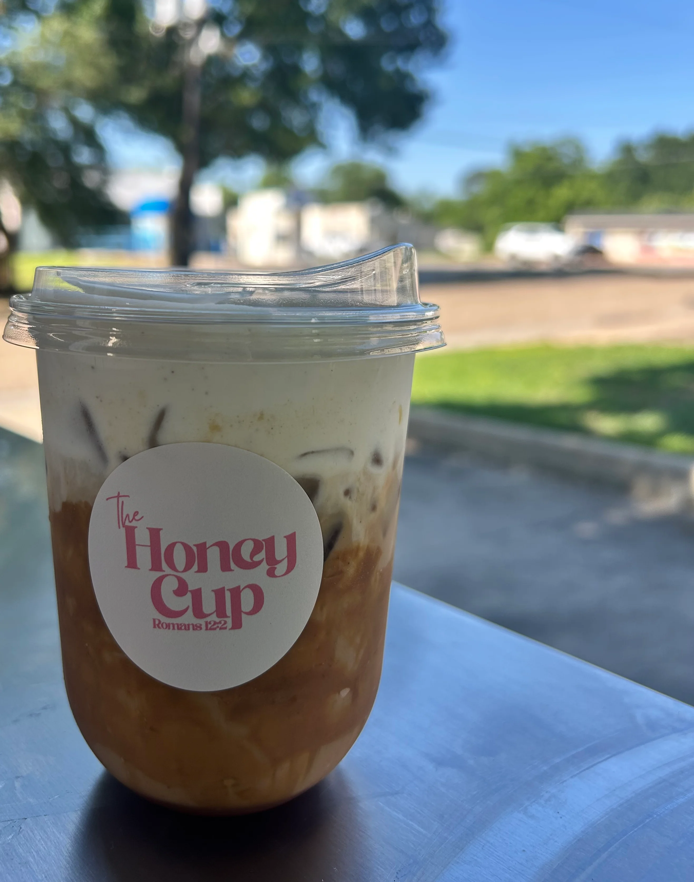 Iced coffee drink from The Honey Cup with a logo and outdoor background.