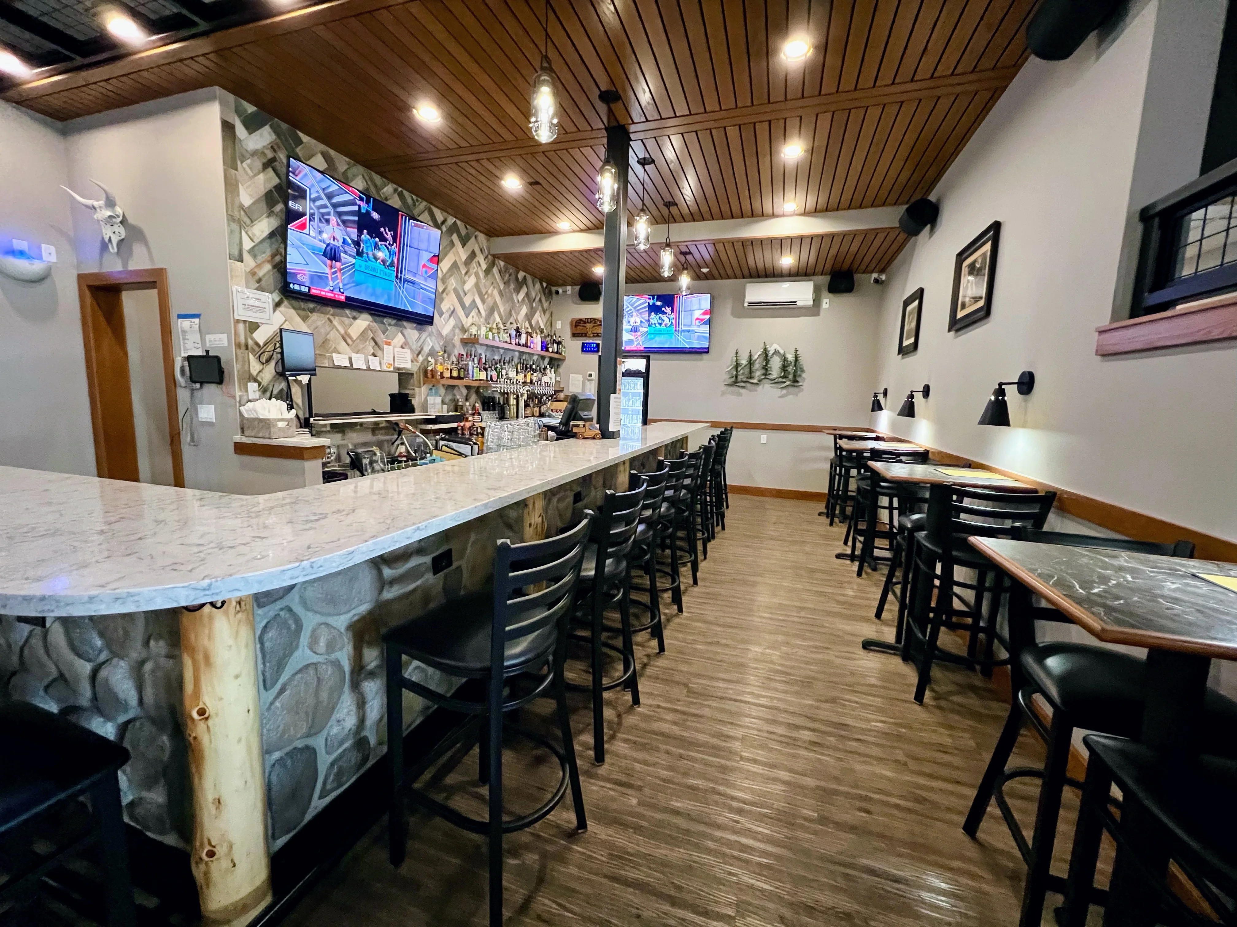 A modern bar area featuring a long stone and marble countertop, with a selection of high-backed chairs and multiple televisions displaying sports.