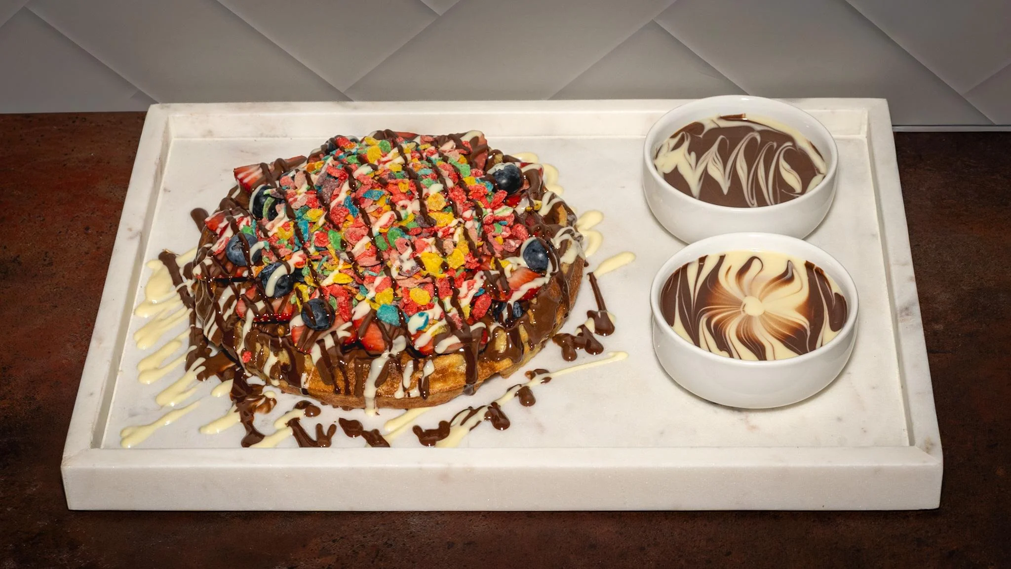 A colorful dessert featuring a waffle topped with various candies and drizzled with chocolate sauce, along with bowls of chocolate and creamy sauce.
