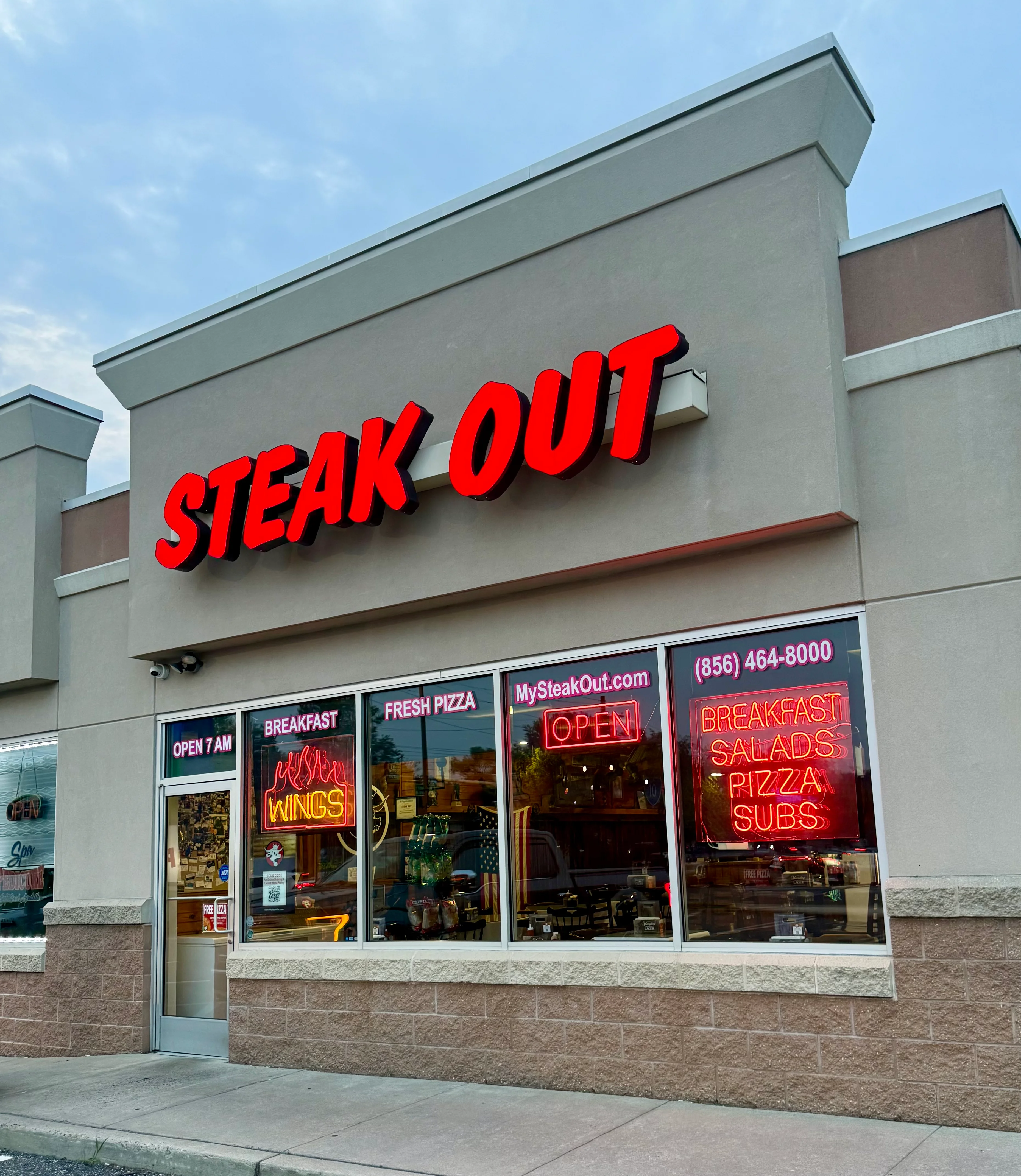 Front entrance of Steak Out in Sewell, NJ with bold red signage and window displays highlighting fresh pizza, wings, salads, and subs