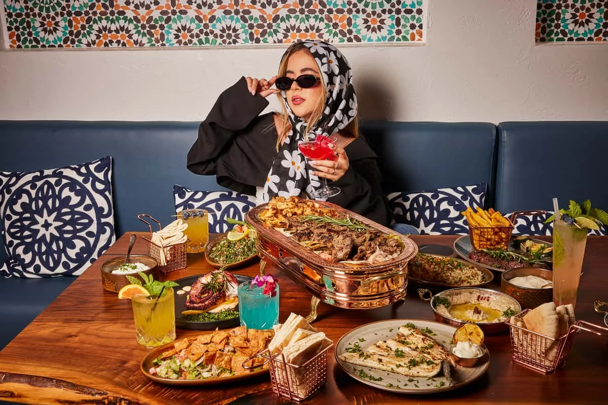 A woman in a floral headscarf and sunglasses holds a red drink at a table with various dishes like grilled meats, pita, and colorful cocktails. The mood is vibrant and lavish.