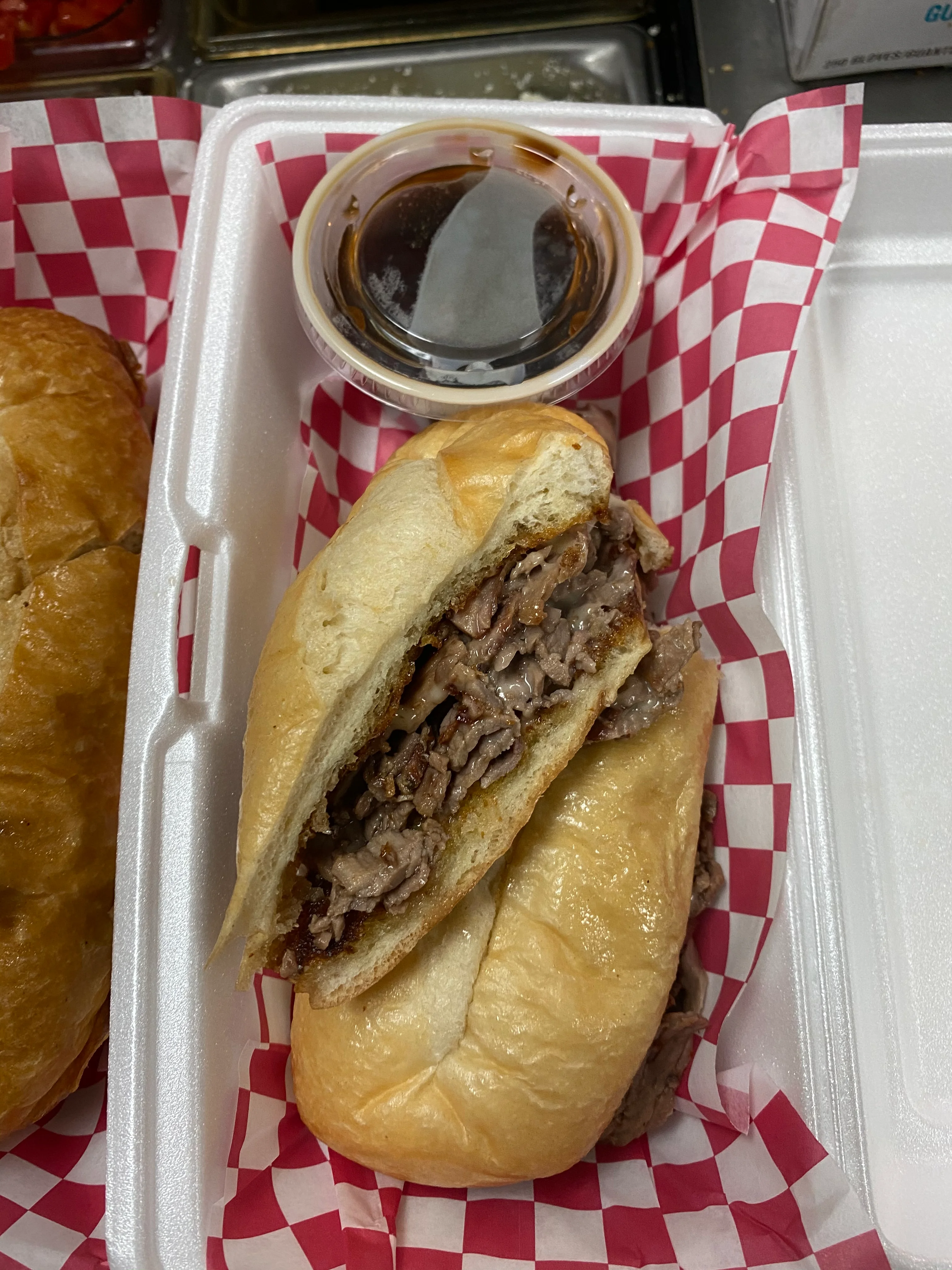 French dip sandwich filled with sliced roast beef on a toasted hoagie roll, served in a red-and-white checkered paper tray with a cup of au jus for dipping.