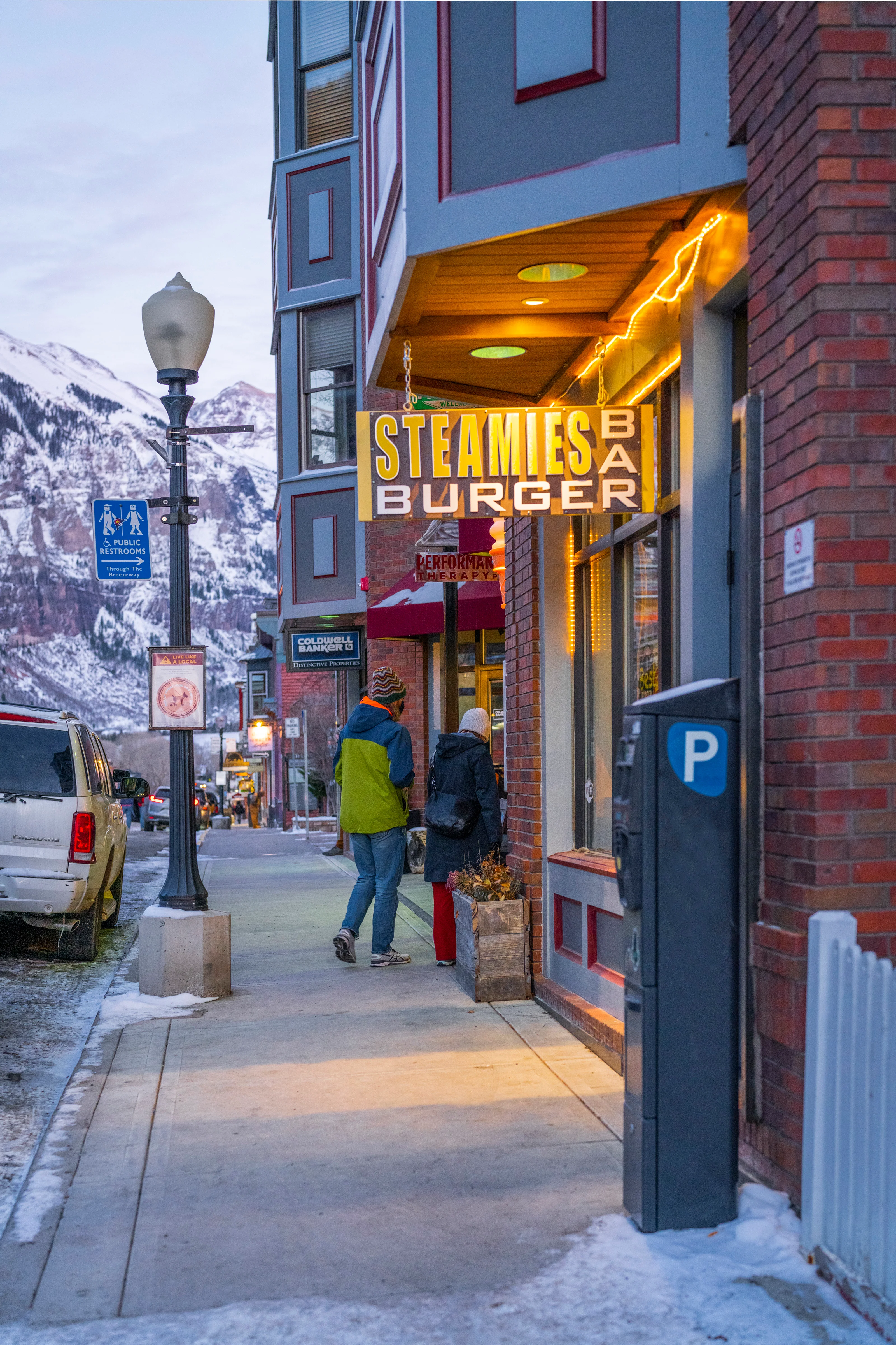 An outdoor view of a burger restaurant with a colorful sign, surrounded by snowy mountains and pedestrians.