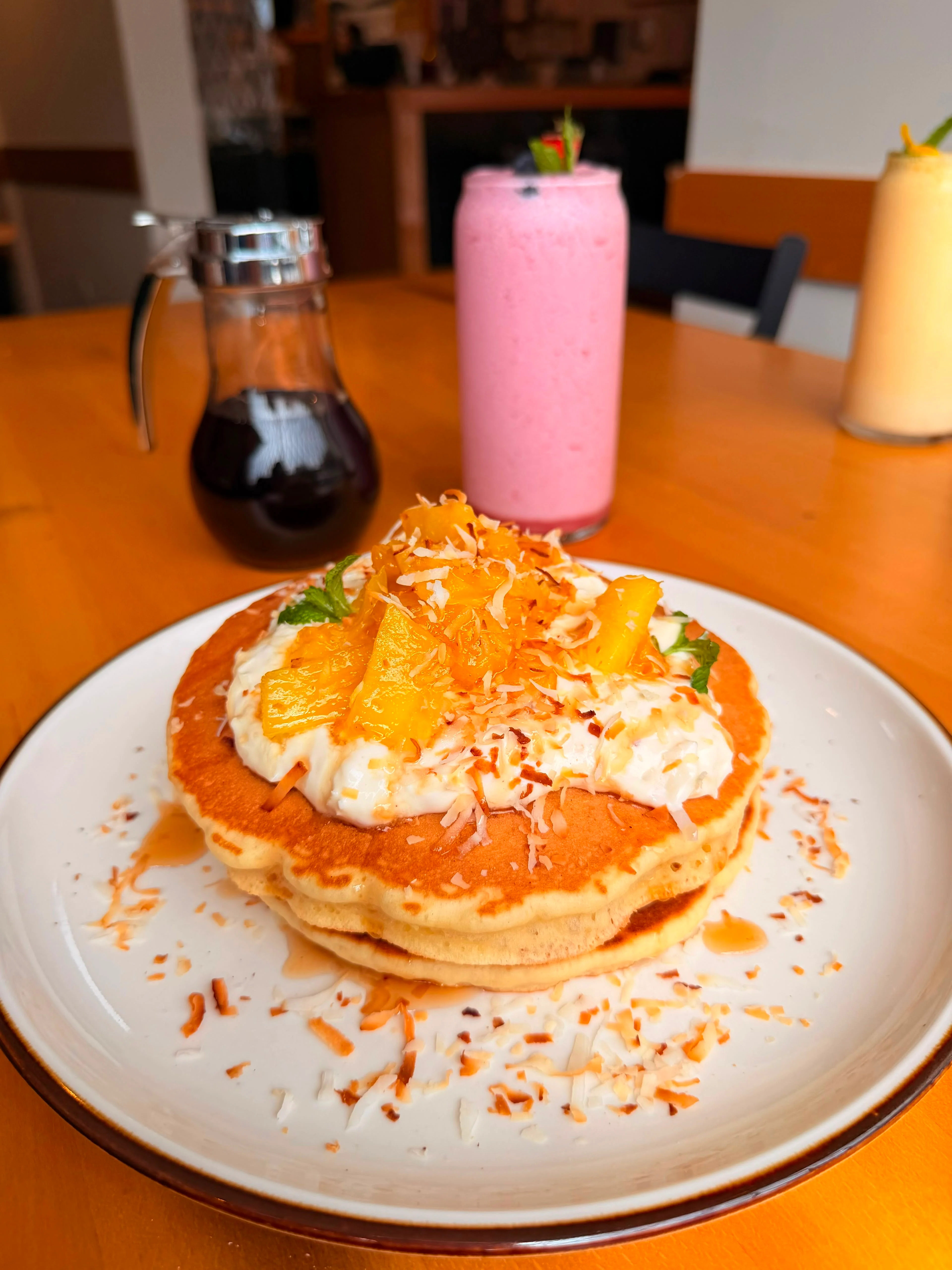 Fluffy pancakes topped with coconut, tropical fruits, and whipped cream, served alongside a sweet smoothie and maple syrup.