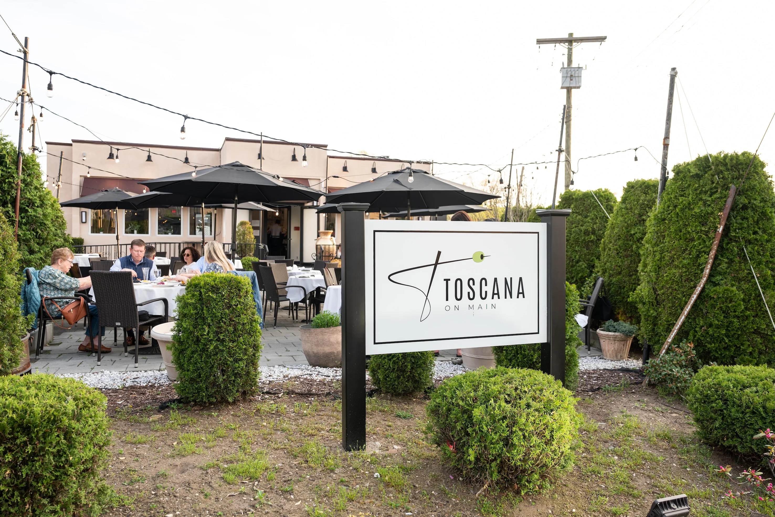 Outdoor seating at "Toscana on Main" restaurant with tables, chairs, black umbrellas, and string lights. 
