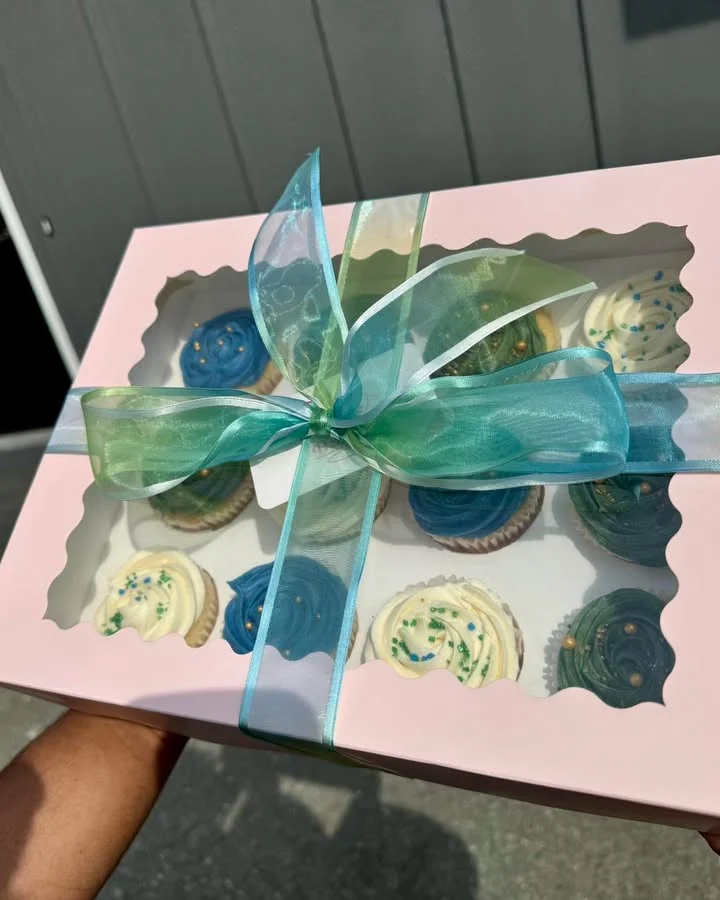A beautifully packaged box of cupcakes, decorated with blue and cream frosting, tied with a ribbon.