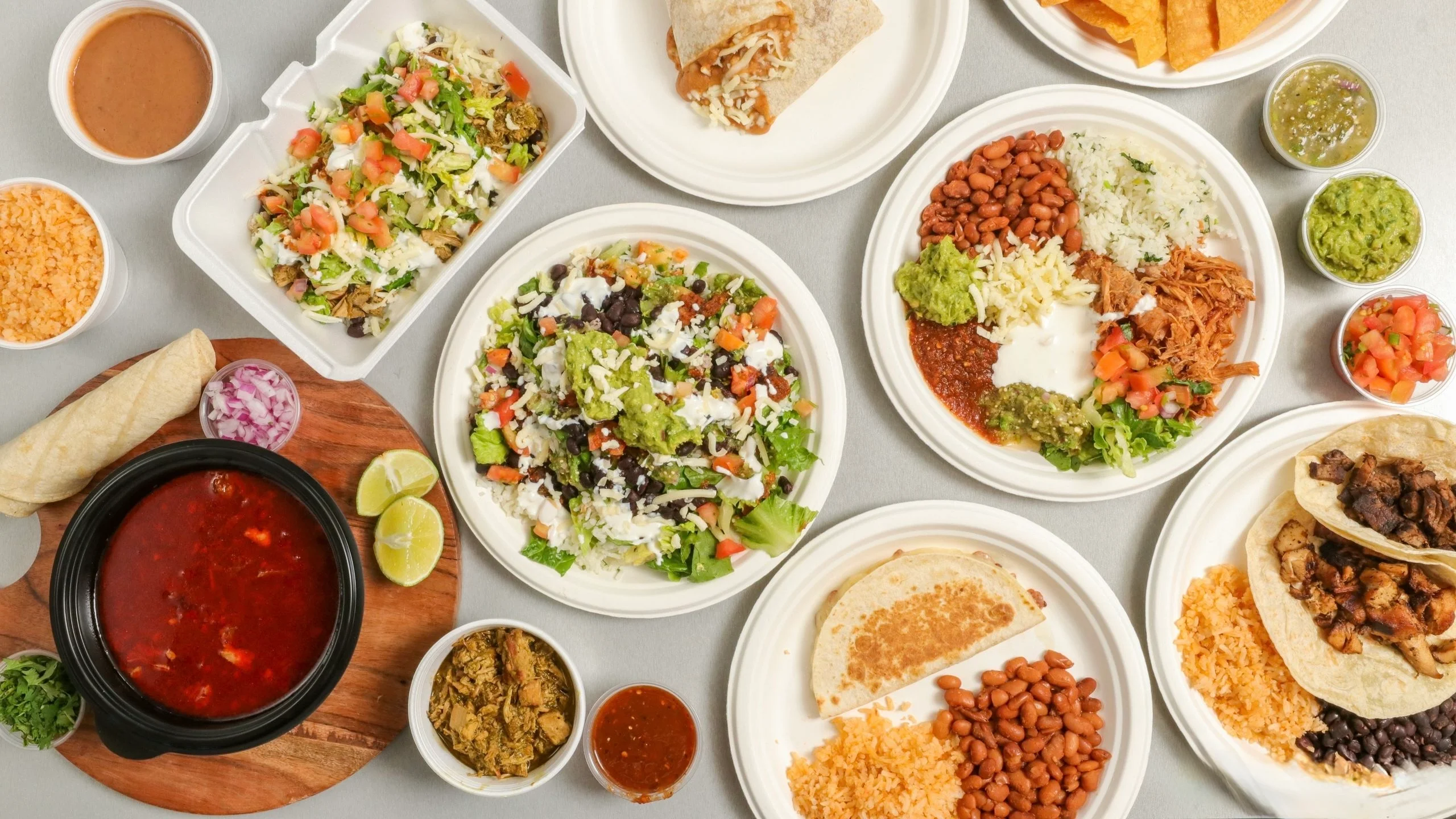 An assortment of Mexican dishes including tacos, burritos, and salads, served on various plates with sides of beans, rice, and sauces.