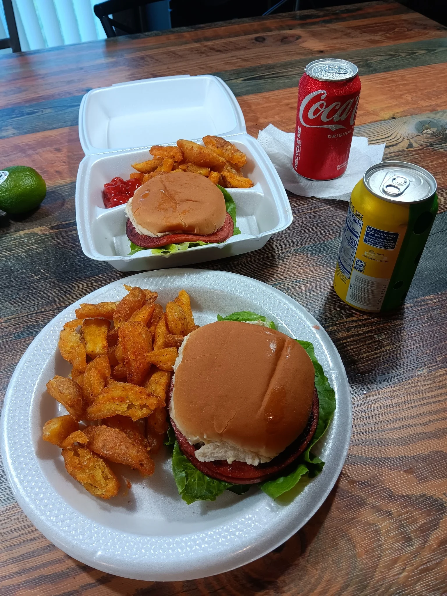 A table setting featuring two hamburgers, seasoned potato wedges, and soft drinks, with a lime in the background.