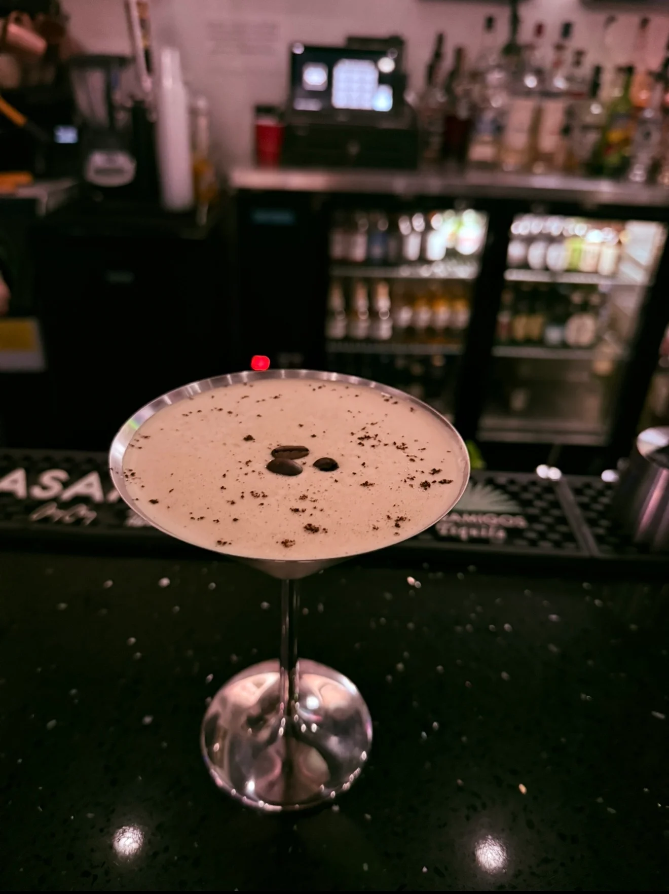 A close-up of a cocktail in a martini glass, topped with coffee beans and a dusting of cocoa, set against a backdrop of bar shelves.