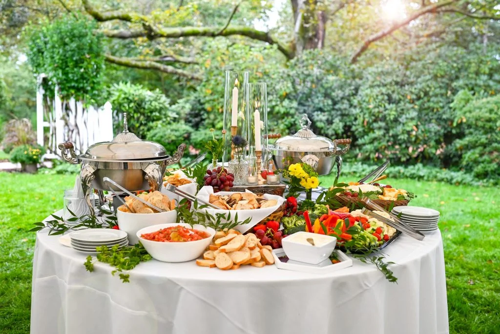 A beautifully arranged outdoor buffet table featuring various dishes, including fruits, breads, and salads, set against a lush green background.