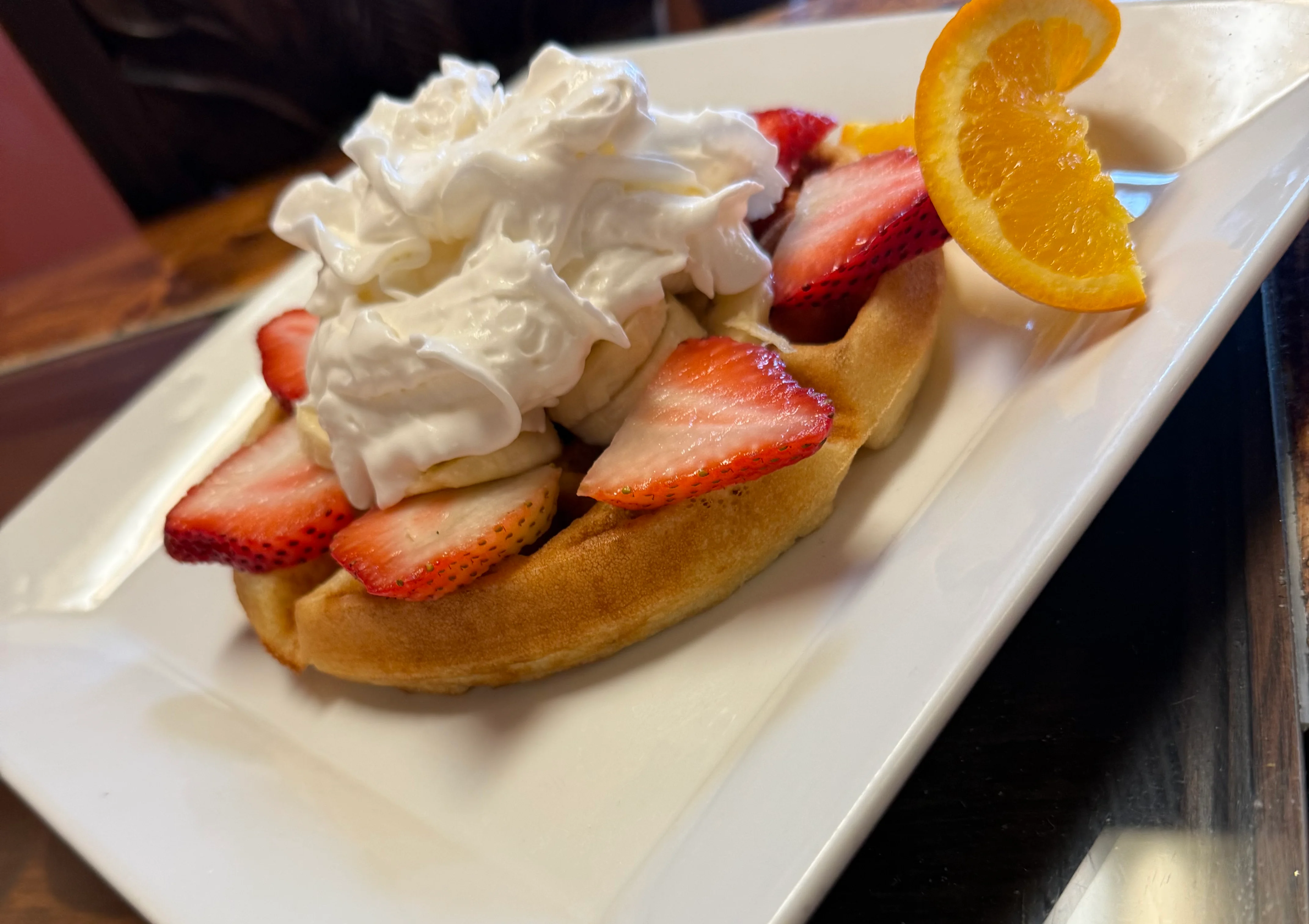 A waffle topped with fresh strawberries and whipped cream, garnished with a slice of orange on a square plate.
