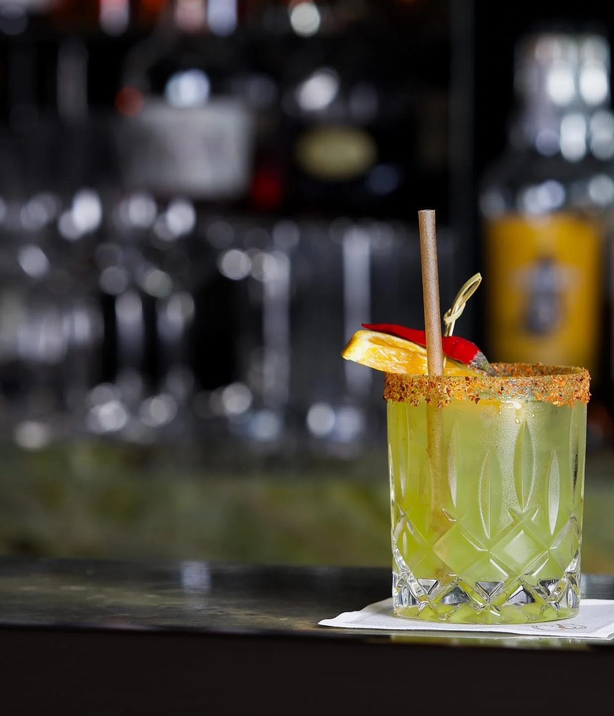 A vibrant green cocktail served in a textured rocks glass with a chili-powder rim, garnished with an orange slice, a red chili pepper, and a straw, resting on a napkin atop a dark bar counter.