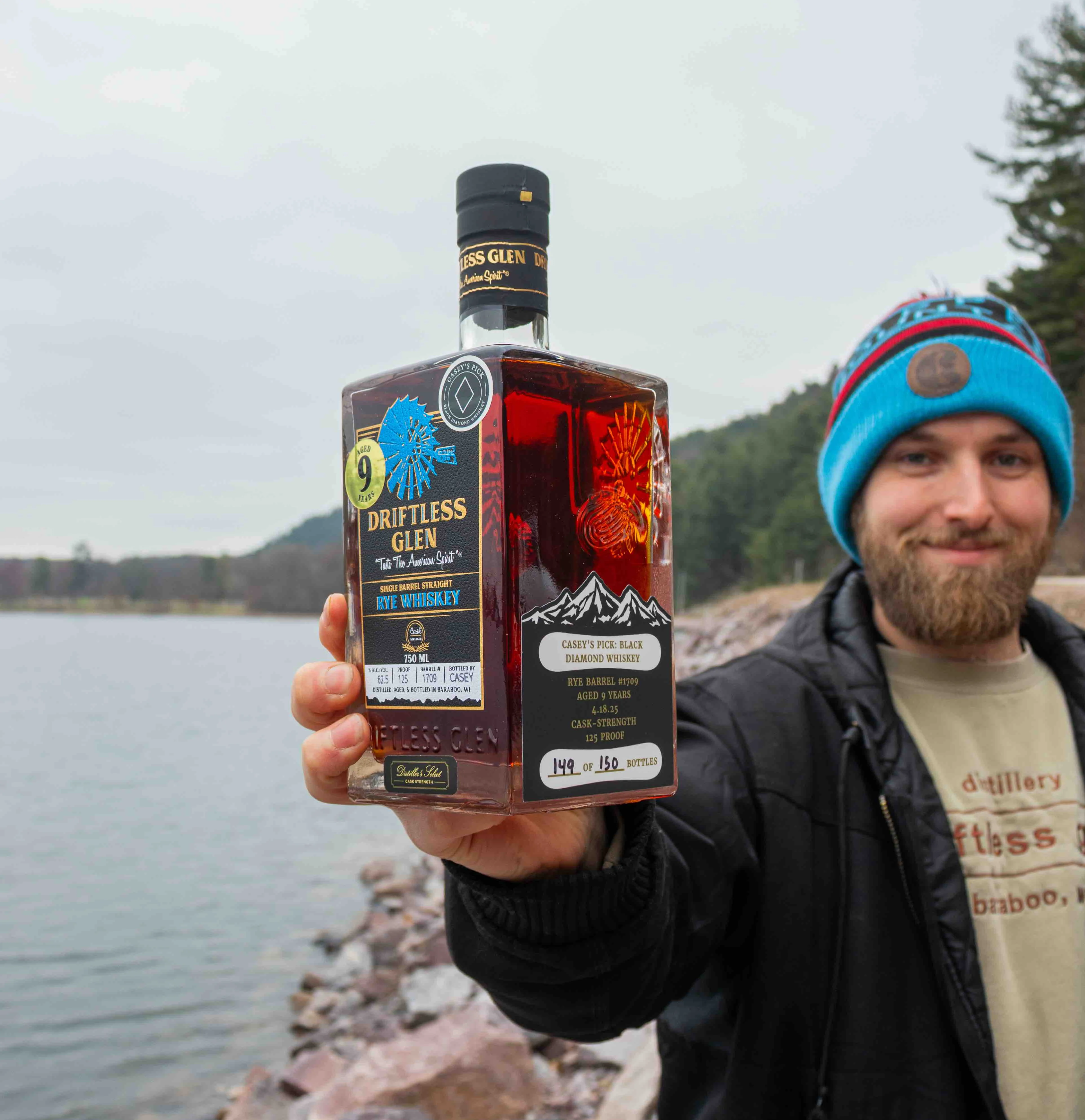 Casey holds a bottle of his Black Diamond Whiskey outside by a lake