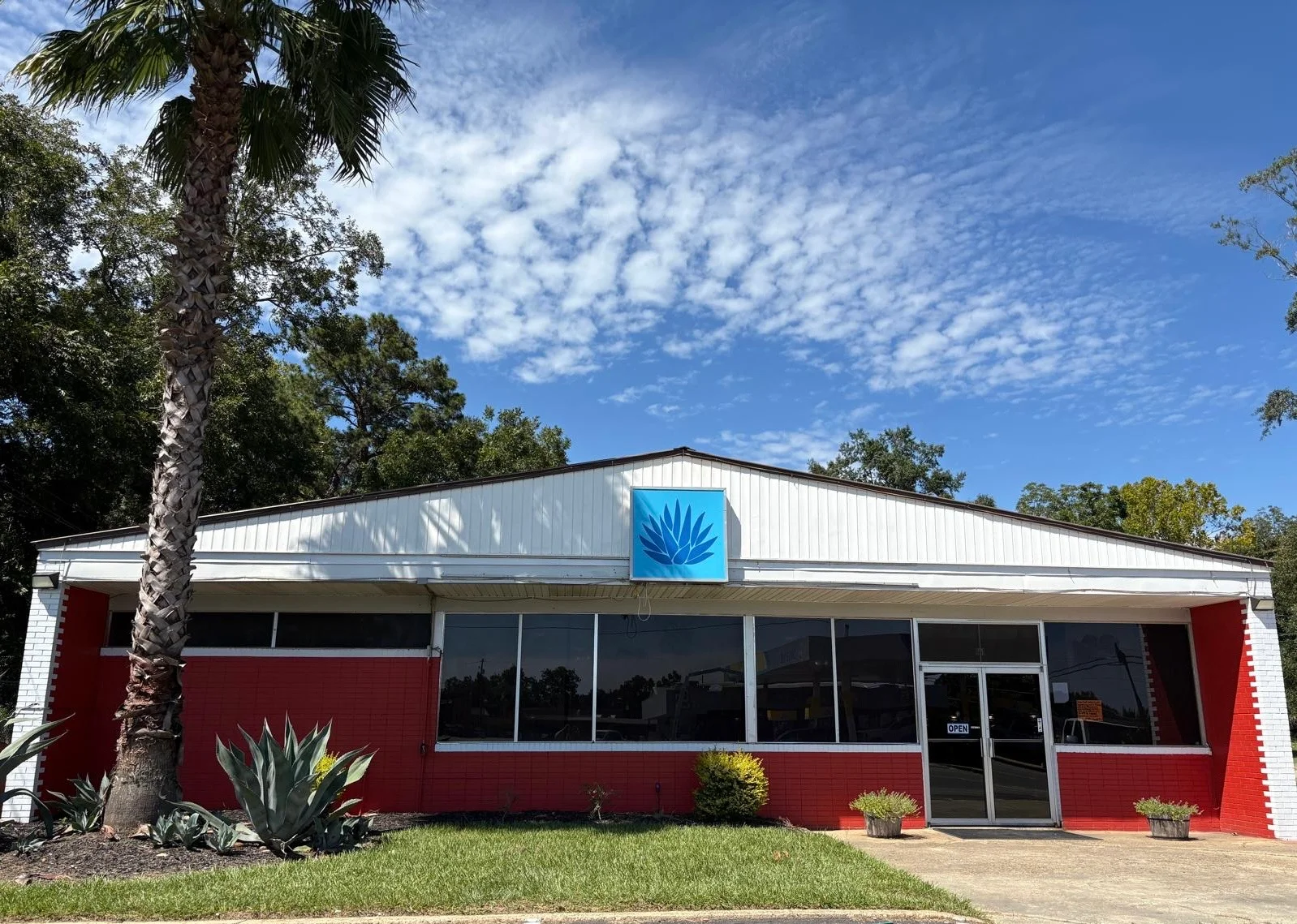 The exterior of a restaurant building with large windows, featuring a blue agave logo on the front and surrounded by landscaping.